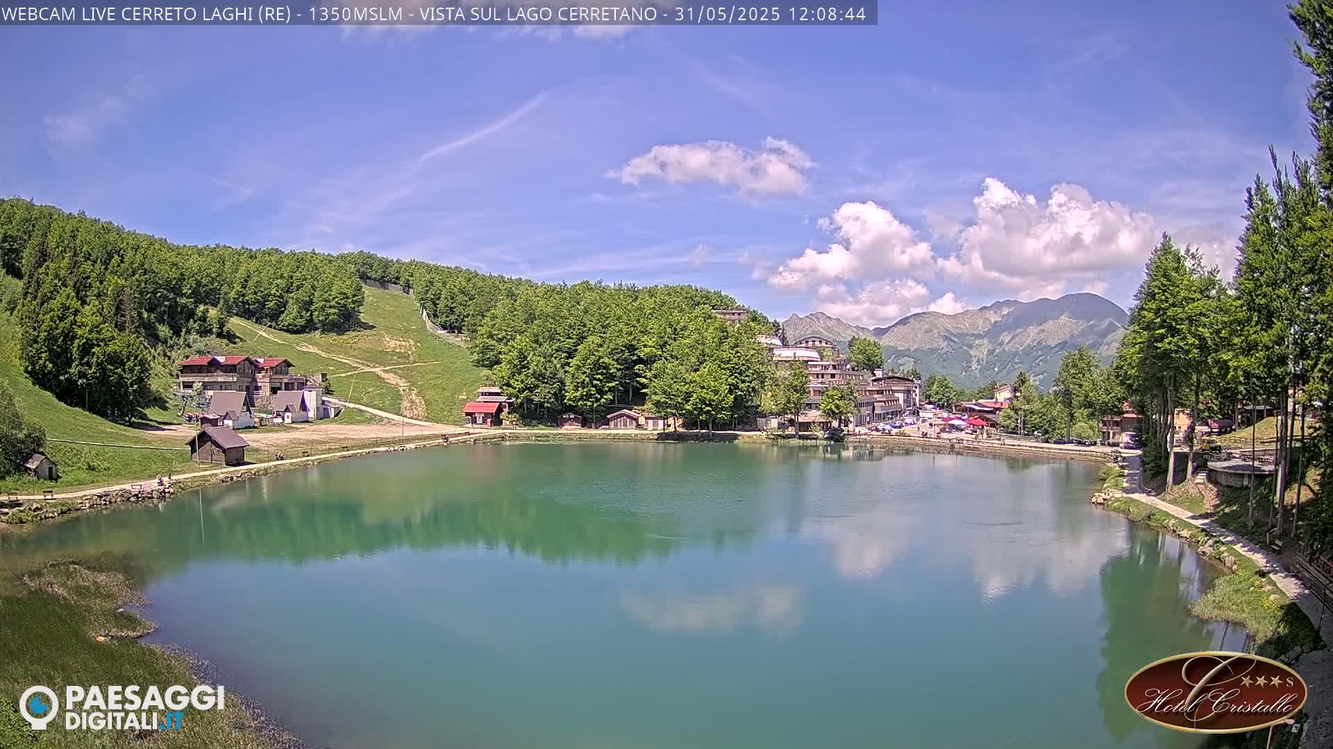 A calm lake reflects a partly cloudy sky and surrounding mountains, with buildings and trees lining the shoreline.