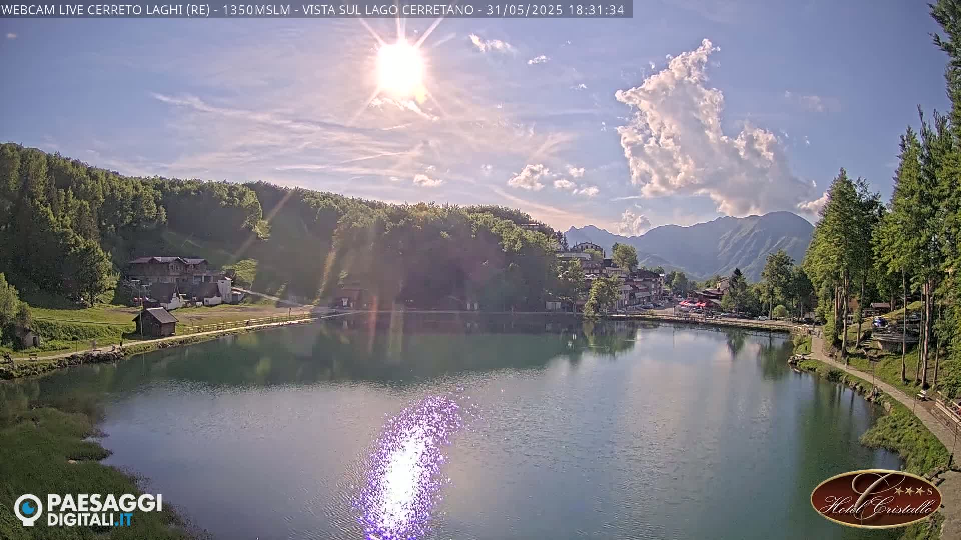 A calm lake reflects the sun's rays, surrounded by green hills, a village, and mountains under a partly cloudy sky.