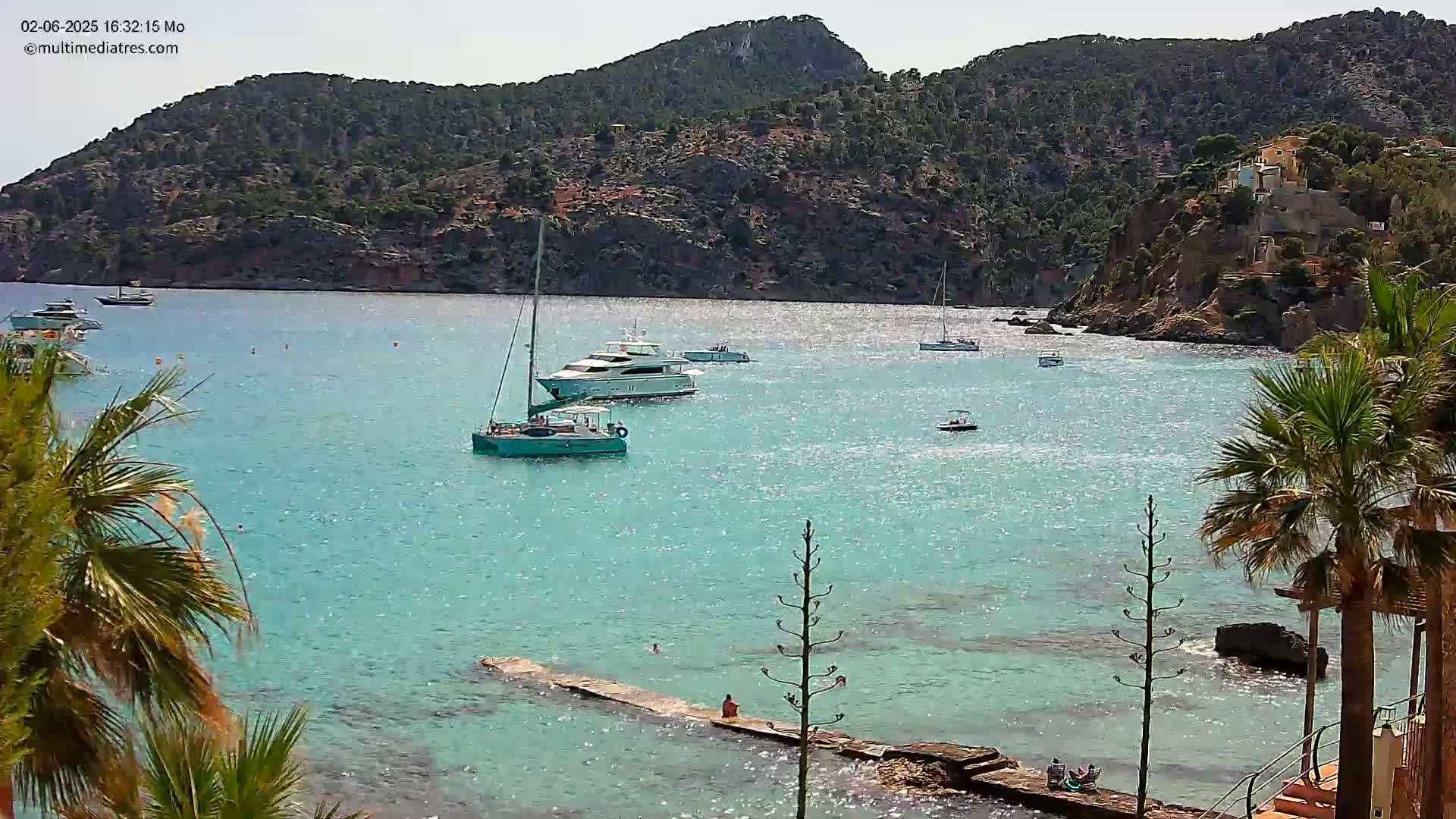 A calm, sunny day shows turquoise water with several boats, a rocky shoreline with houses on the hills, and palm trees in the foreground.