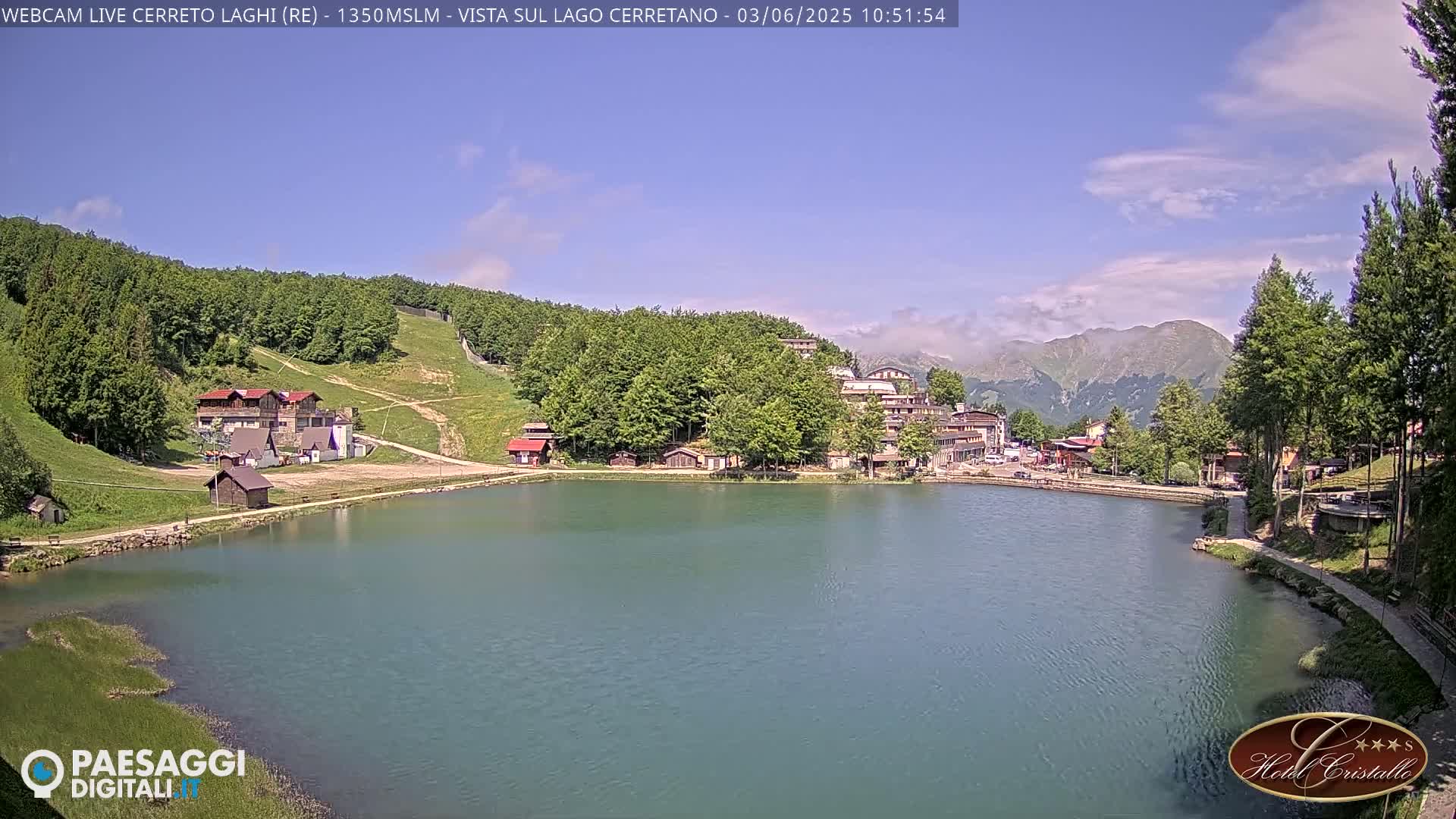 A calm lake is nestled amongst a green, hilly landscape under a mostly sunny sky with a few clouds.