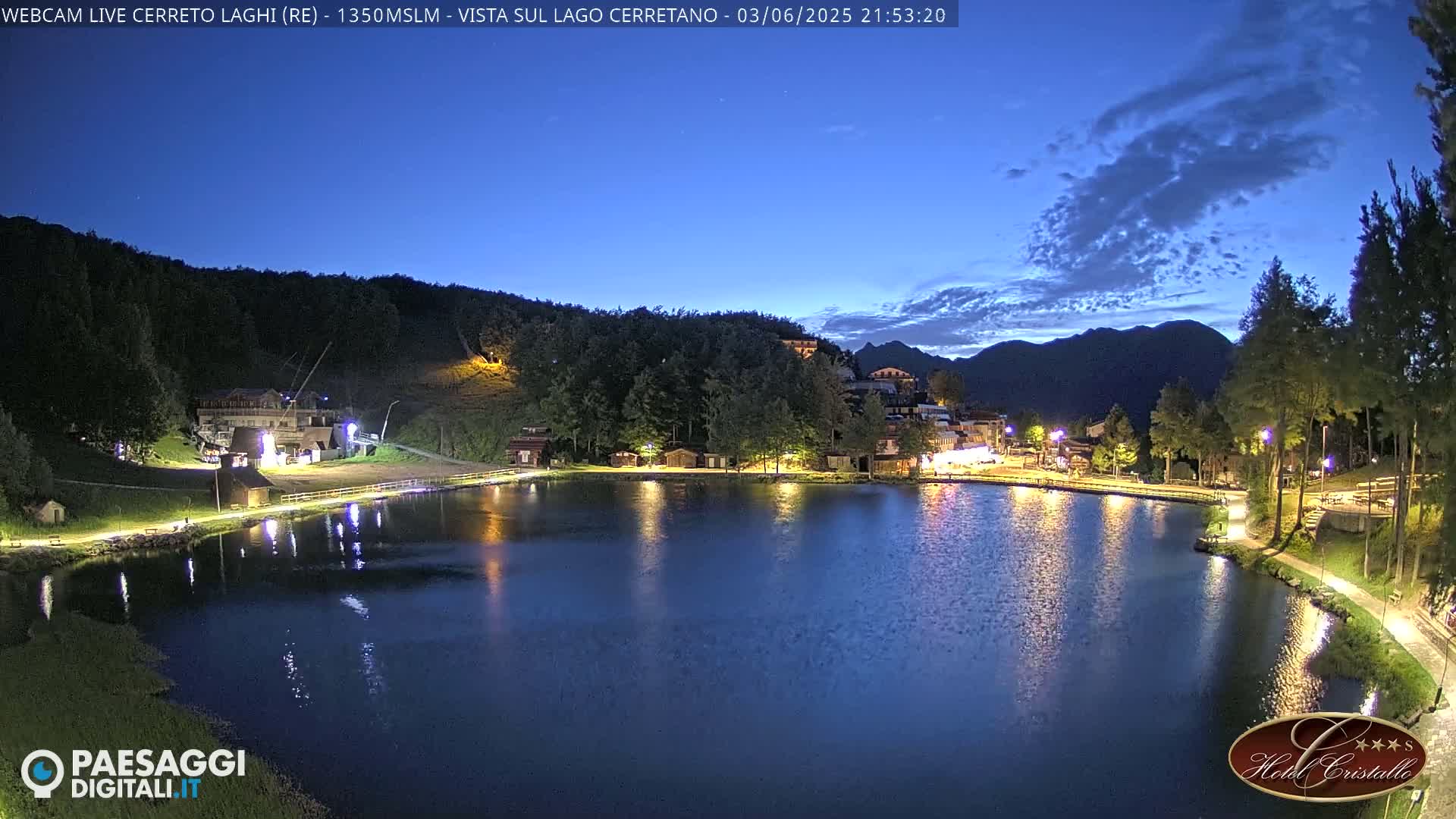 A tranquil lake at twilight reflects the lights of a small village nestled against a dark, tree-covered hillside under a mostly clear, dark blue sky.