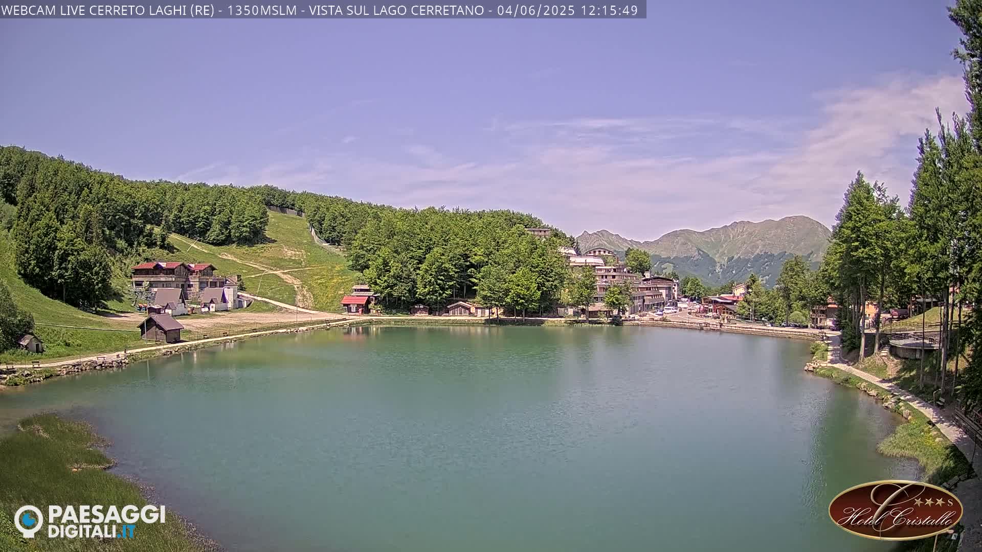 A calm lake is nestled in a mountainous region under a mostly clear, sunny sky, with buildings and greenery surrounding its shores.