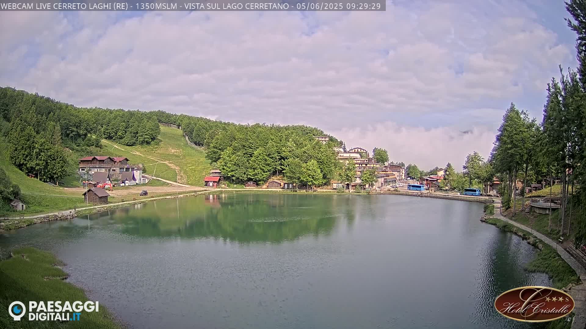 A calm lake reflects the surrounding buildings and lush green hills under a partly cloudy sky.