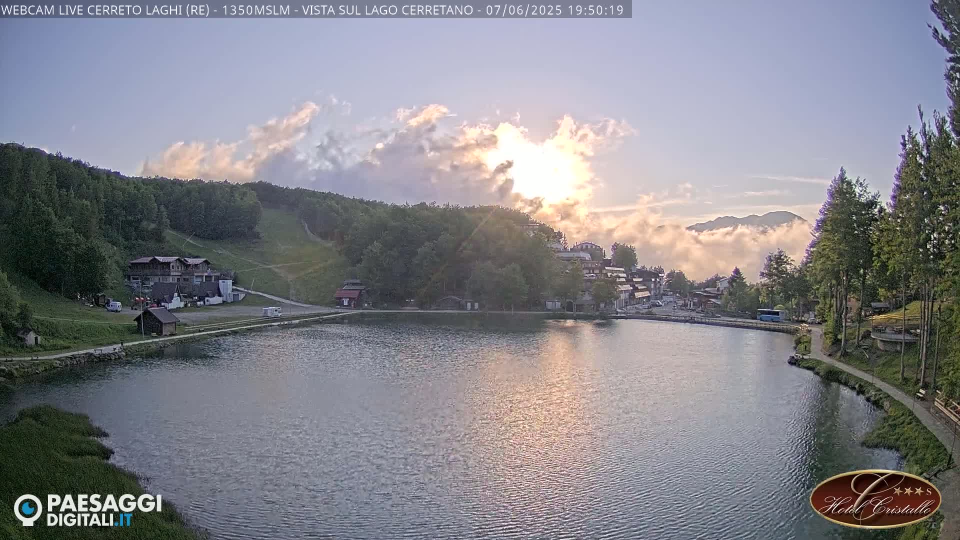 A calm lake reflects a sunset, partially obscured by clouds, with a village and forested hills in the background.