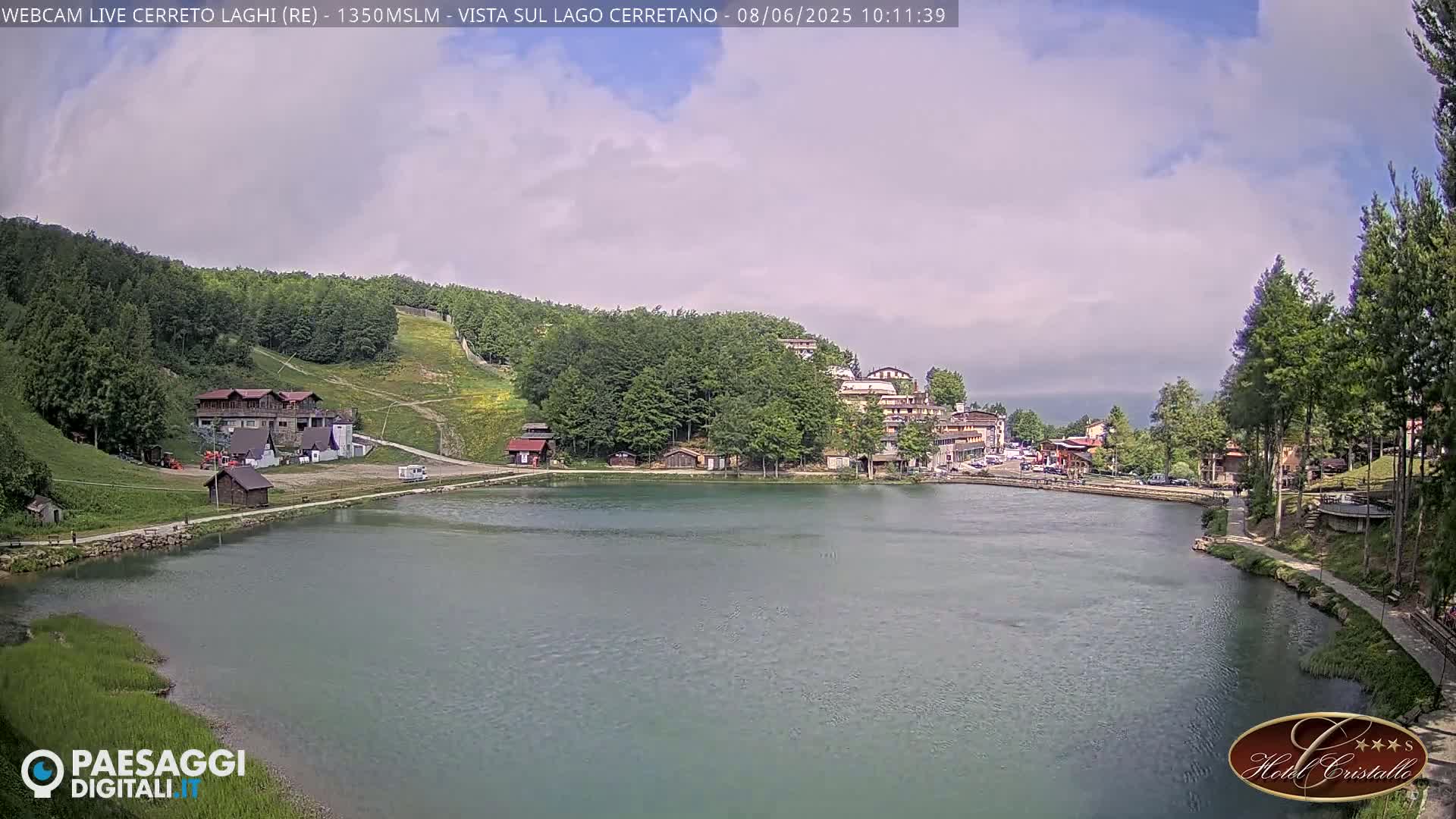 A calm lake is nestled among a green, hilly landscape with buildings and trees under a partly cloudy sky.