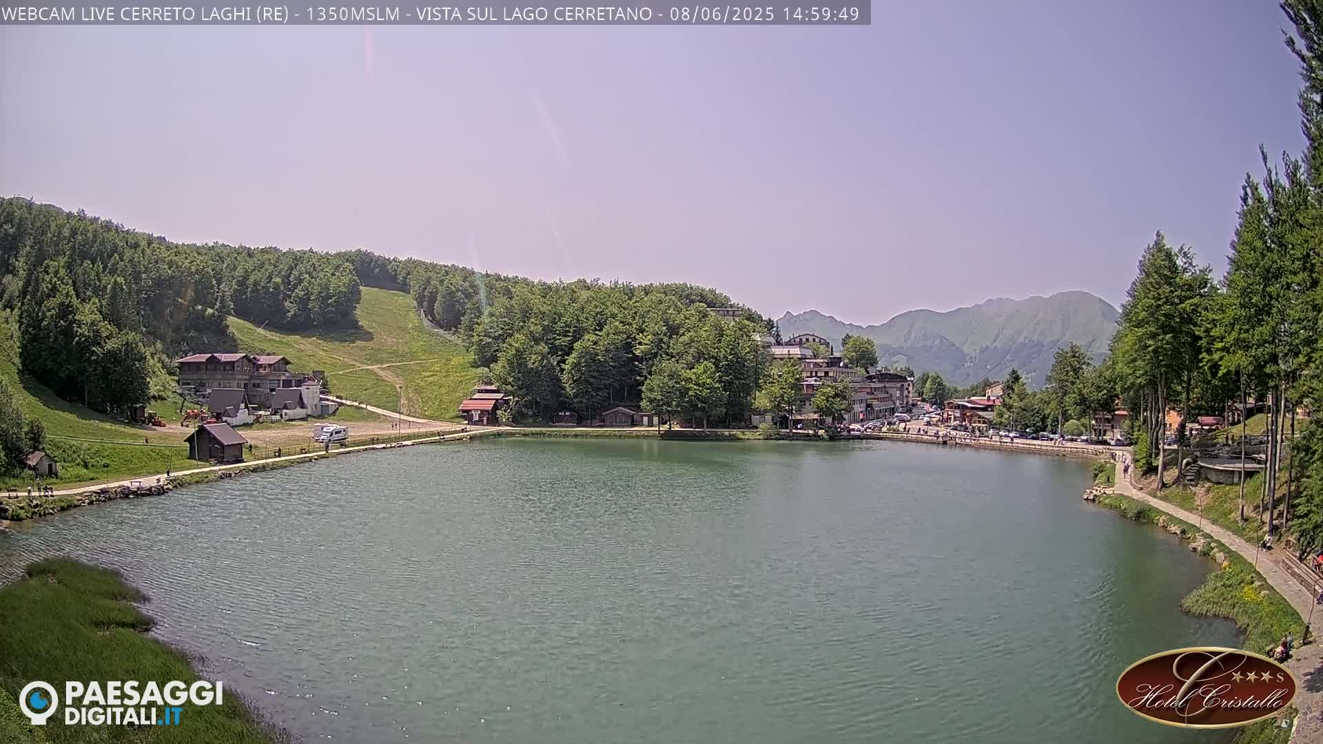 A calm lake is nestled between a mountainside ski slope and a village, under a clear, sunny sky.