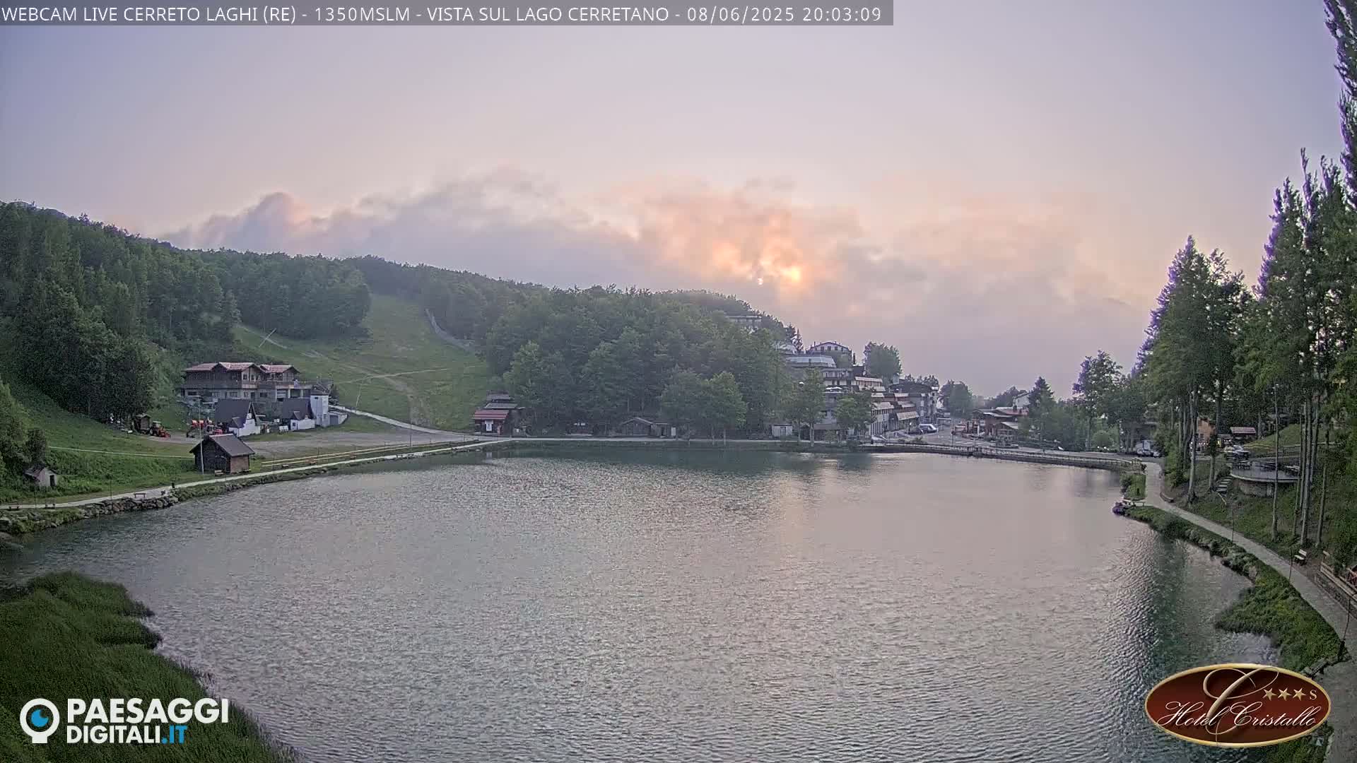 A calm lake sits below a village nestled in a hillside forest, under a hazy, pinkish sunset sky.
