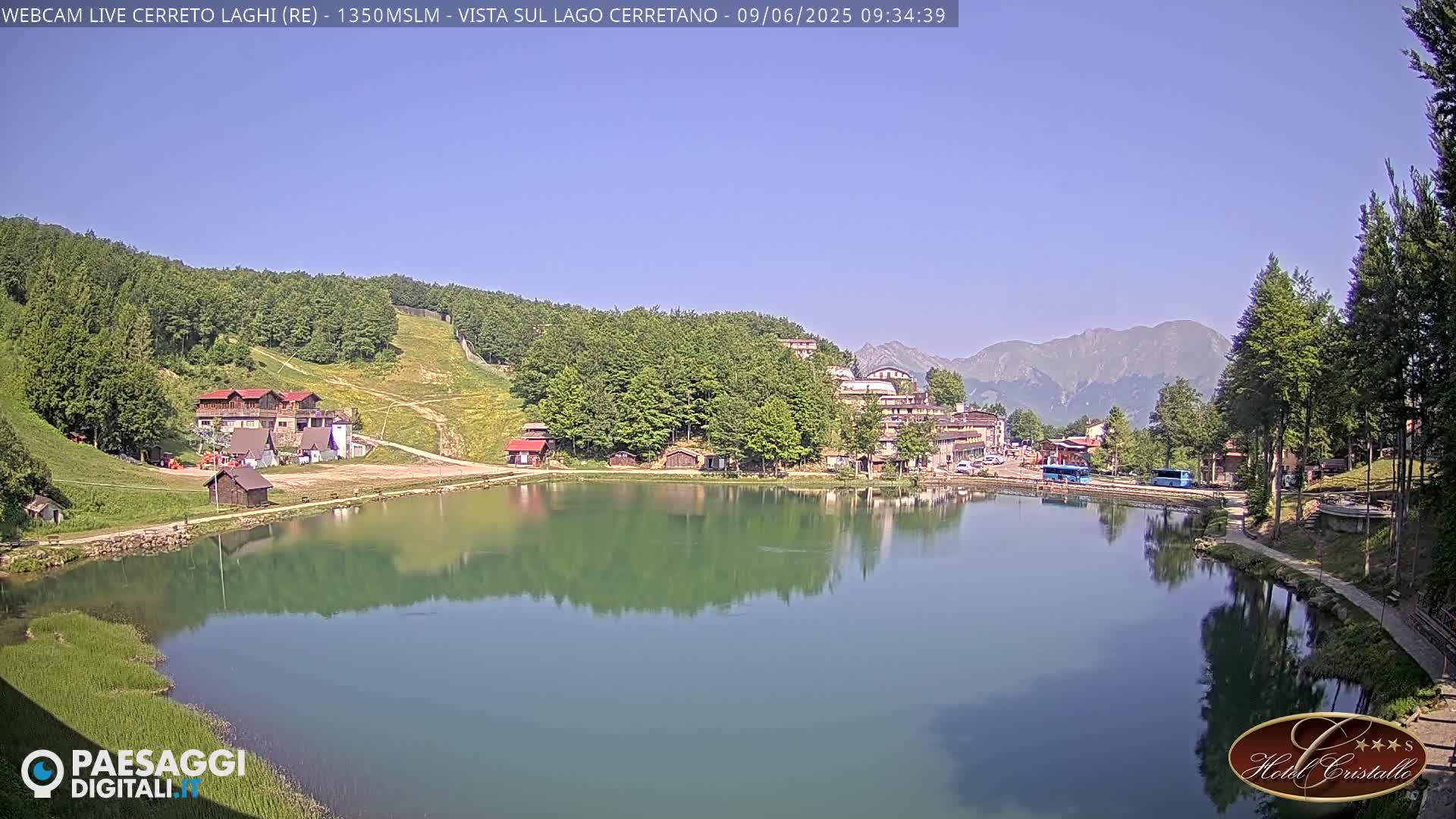 A calm lake reflects a mountain village nestled among green hills and mountains under a clear blue sky.