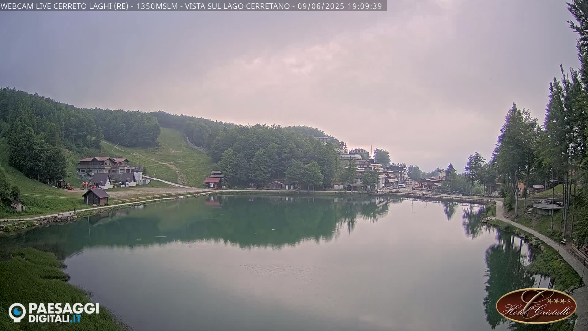 A calm lake reflects a village nestled in a hillside of green trees under an overcast sky.
