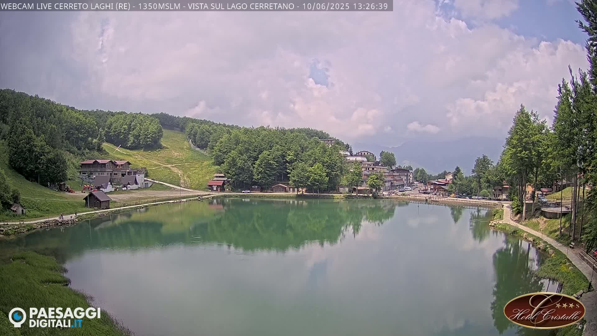 A calm lake reflects the surrounding mountain scenery, which includes buildings and a ski slope, under a partly cloudy sky.