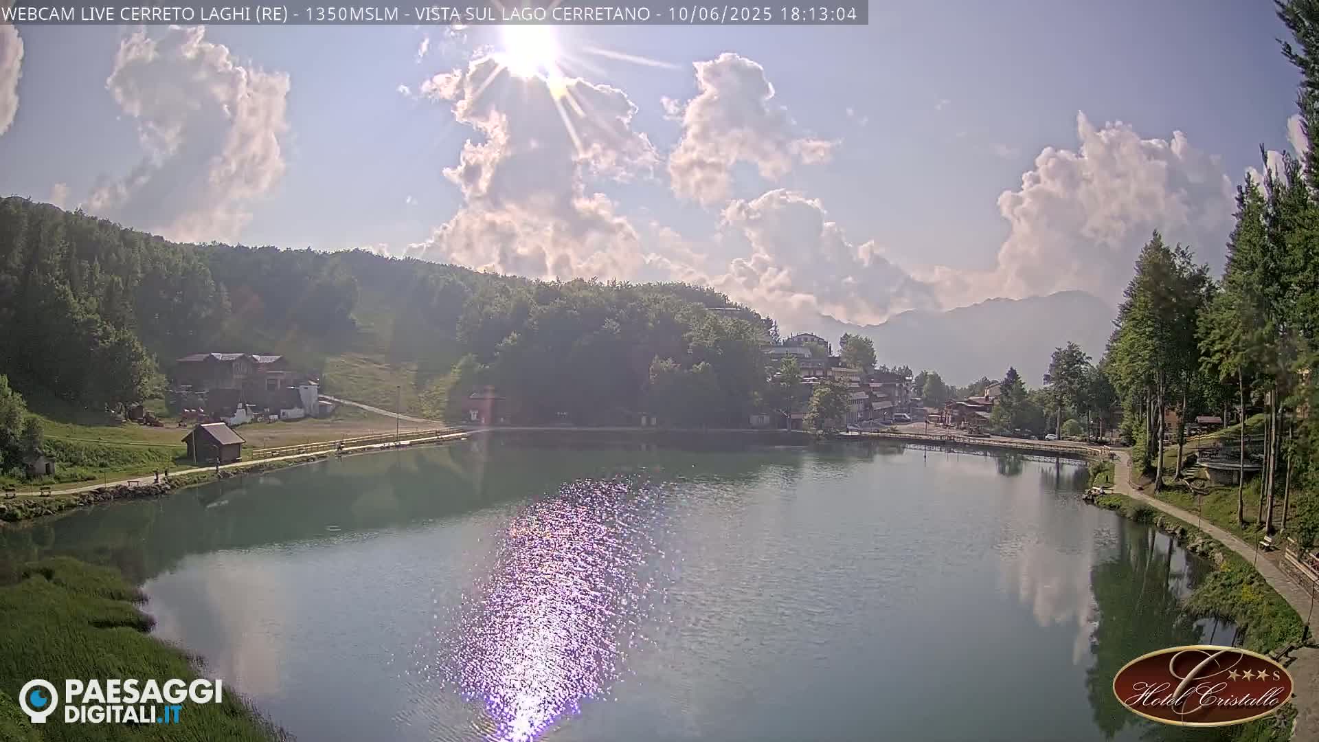 A calm lake reflects the sunlight and clouds in a partly sunny sky, nestled amongst green hills and a small village.