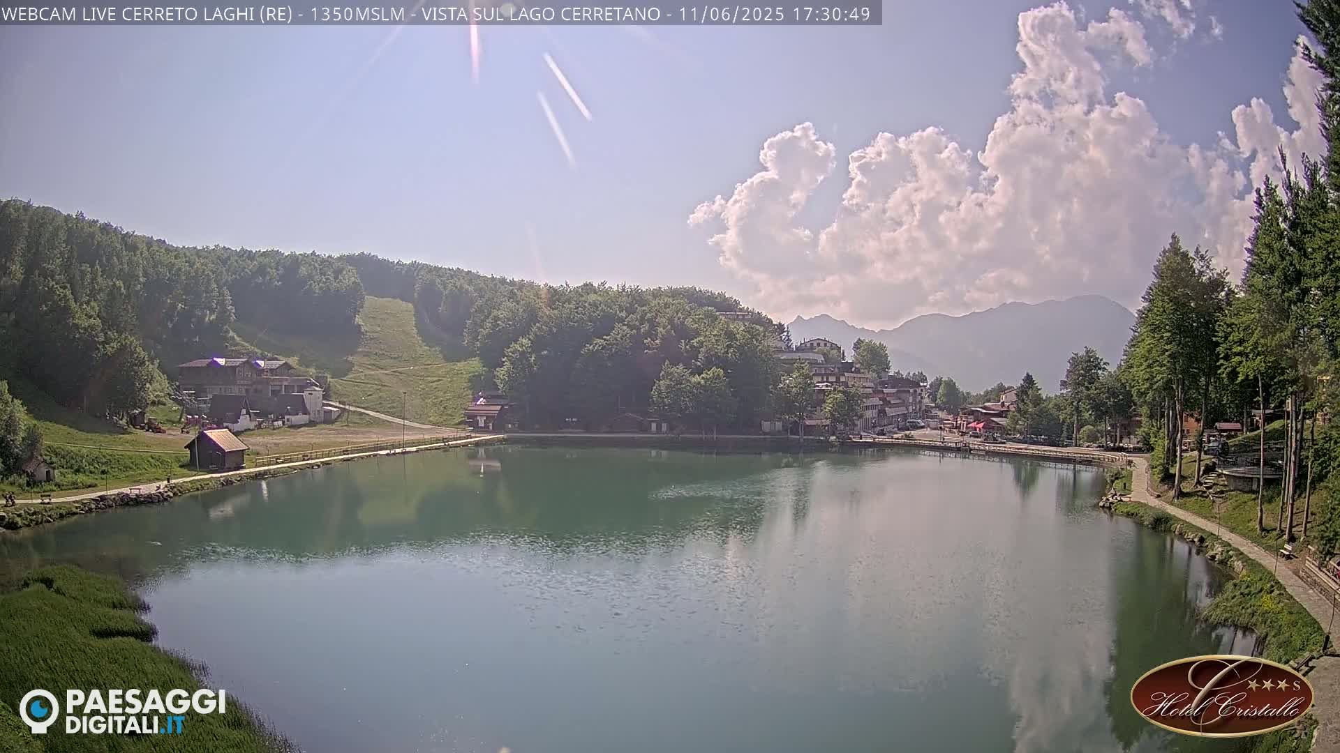 A partly cloudy day reveals a calm lake nestled in a valley surrounded by green hills, buildings, and trees, with mountains visible in the distance.