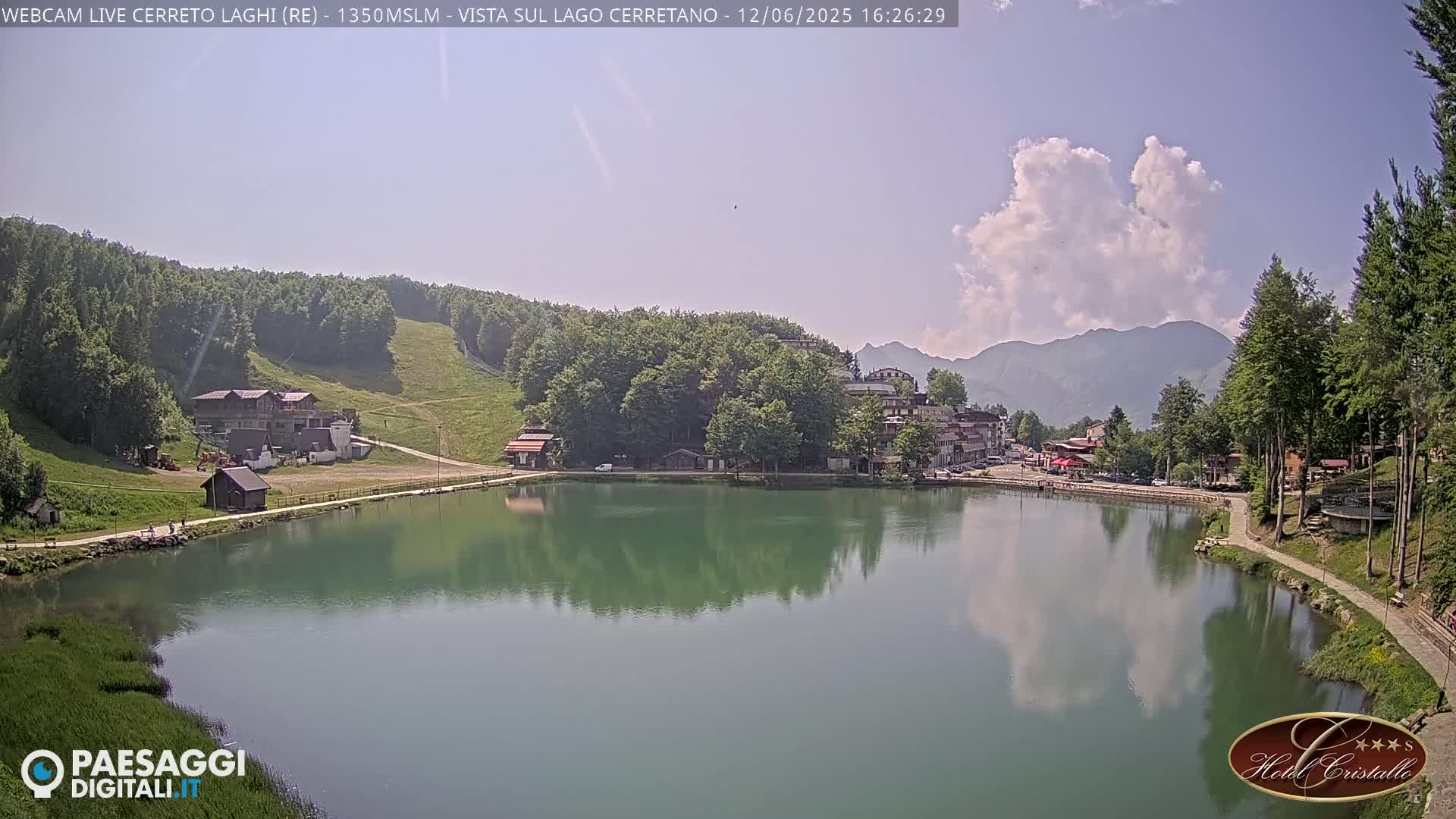A calm lake reflects the surrounding mountains and buildings under a partly cloudy sky.