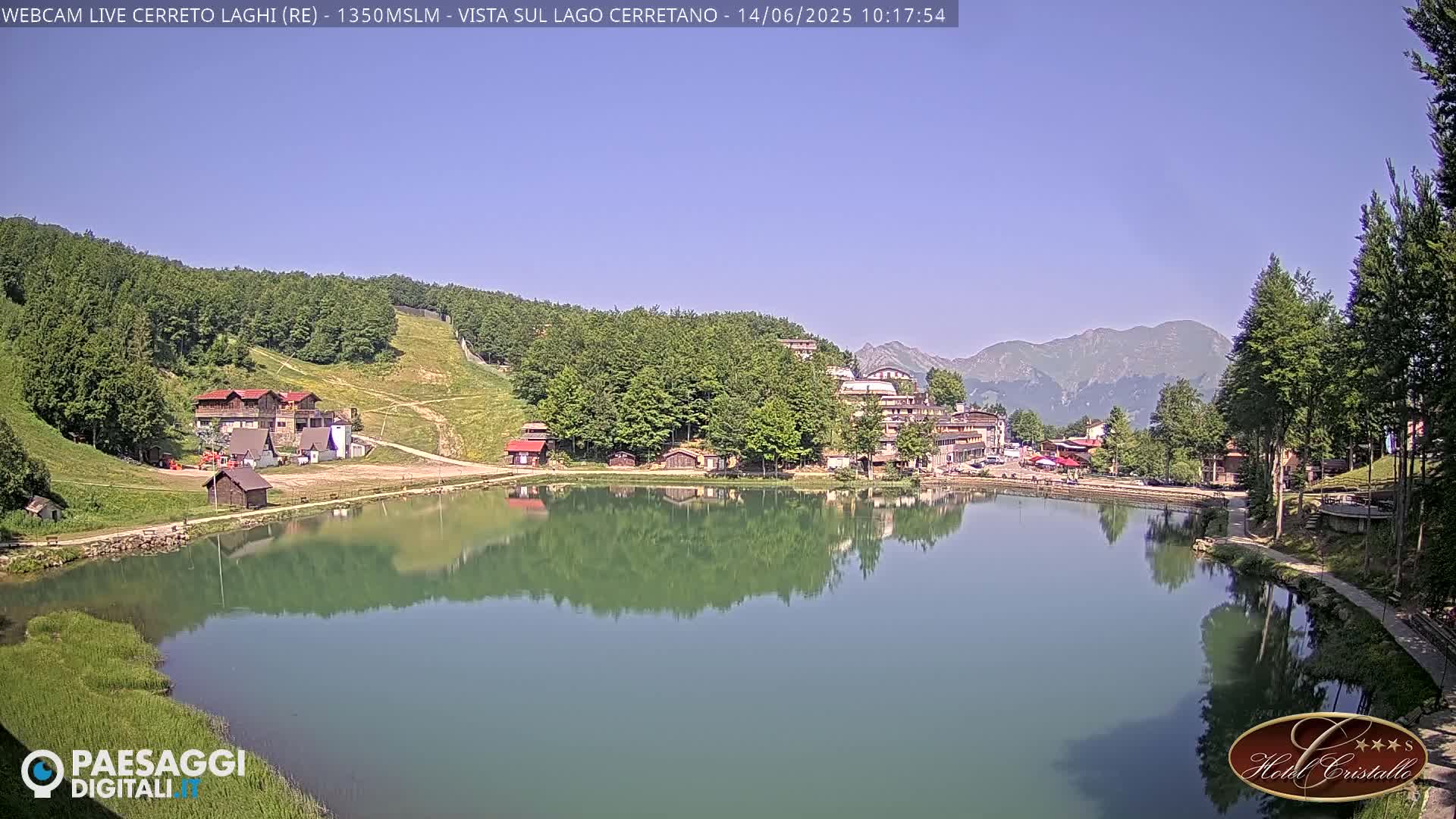 A calm lake reflects a small village nestled in a mountain valley under a clear, sunny sky.
