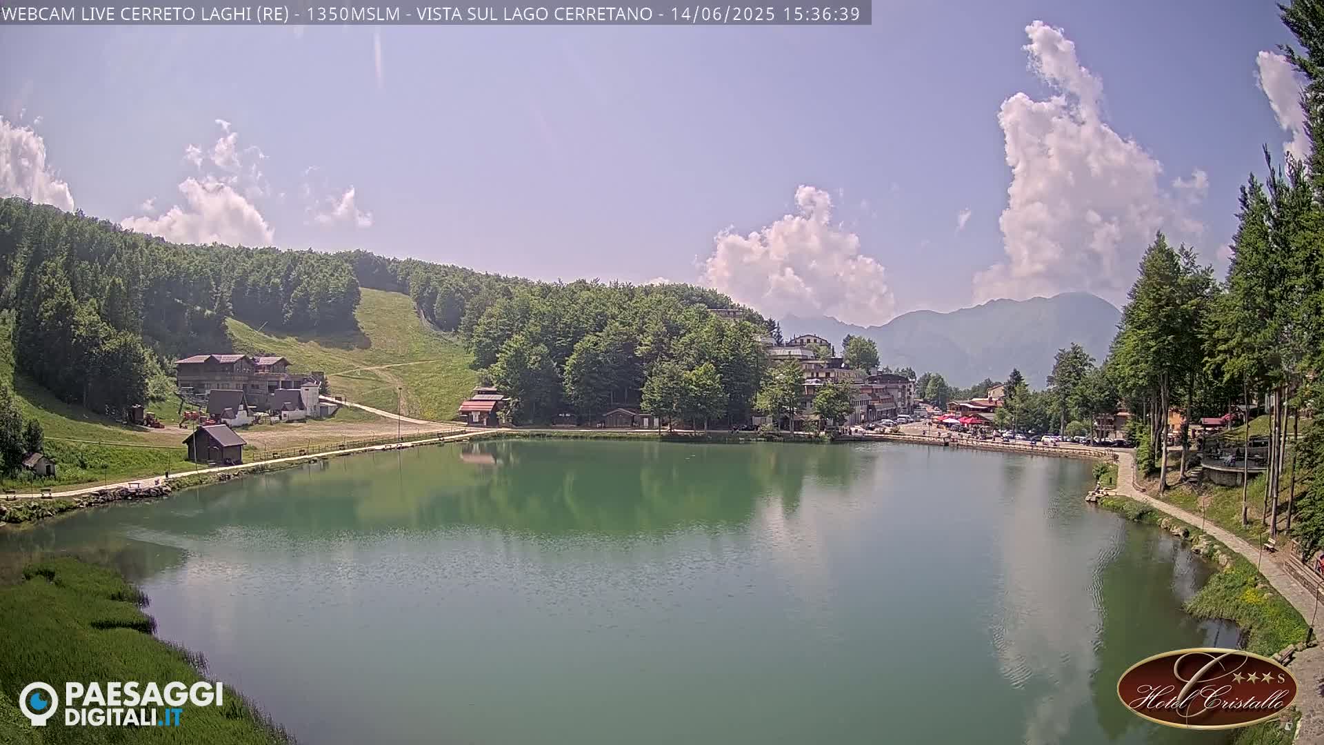 A calm, green lake is surrounded by a village nestled in a mountain valley under a partly cloudy sky.