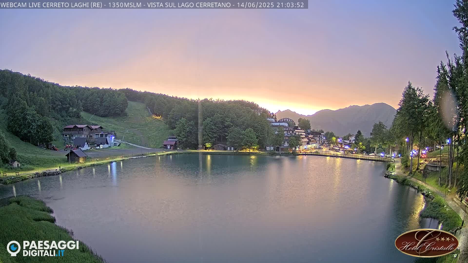 A calm, green lake is surrounded by a village nestled in a mountain valley under a partly cloudy sky.
