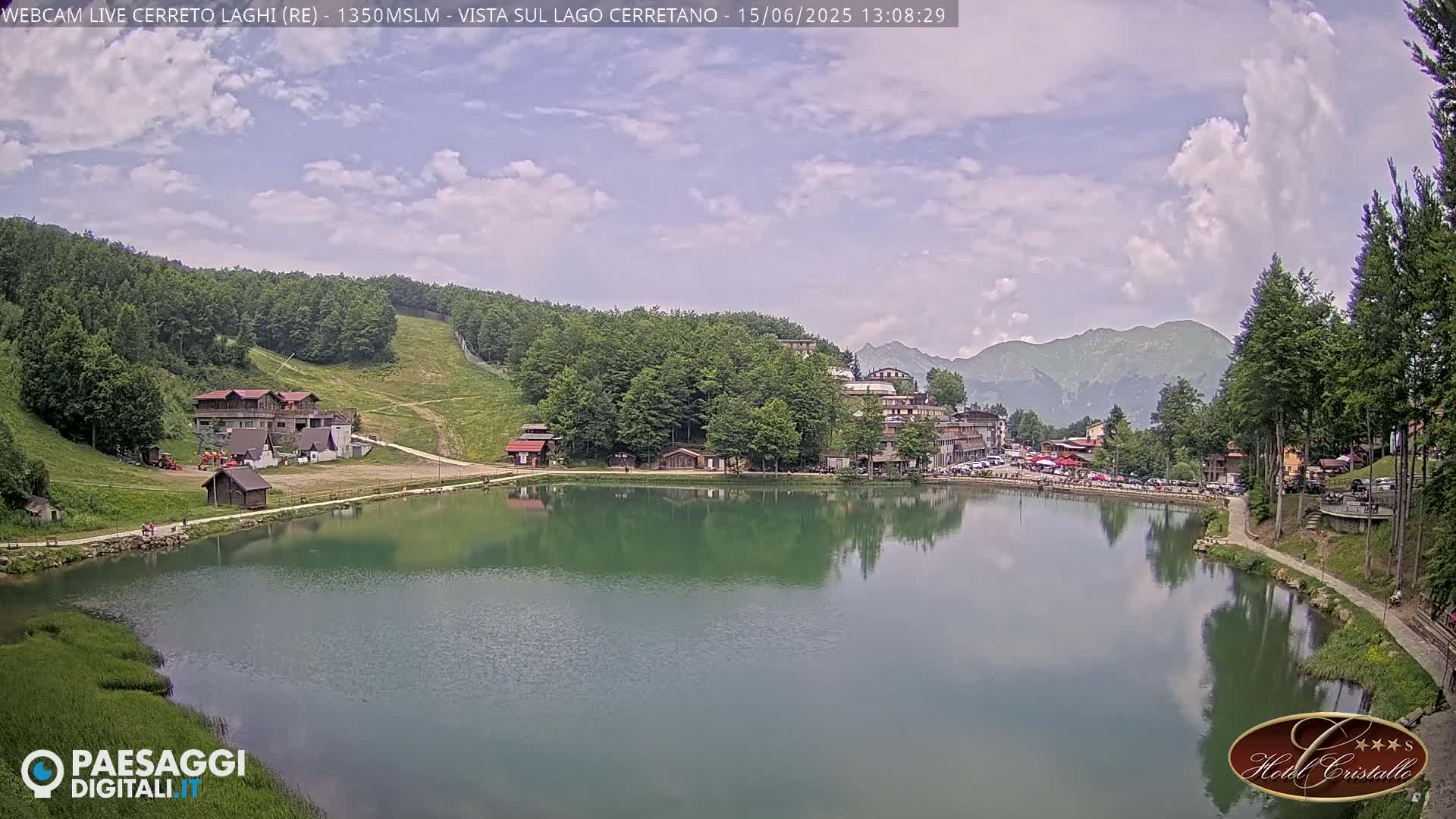 A calm lake reflects a village nestled in a mountain valley under a partly cloudy sky.