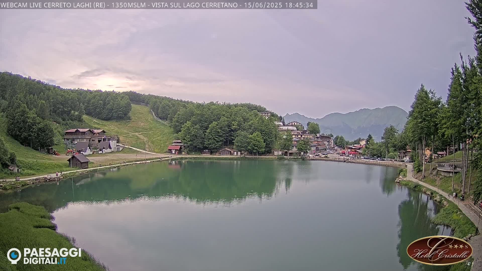 A calm lake reflects a village nestled among green hills under a cloudy, overcast sky.