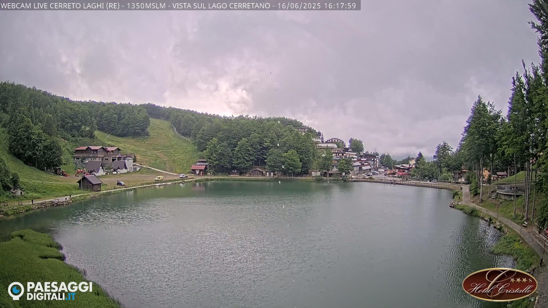 A calm lake sits below a hillside village under an overcast sky, with green trees and a ski slope visible in the background.