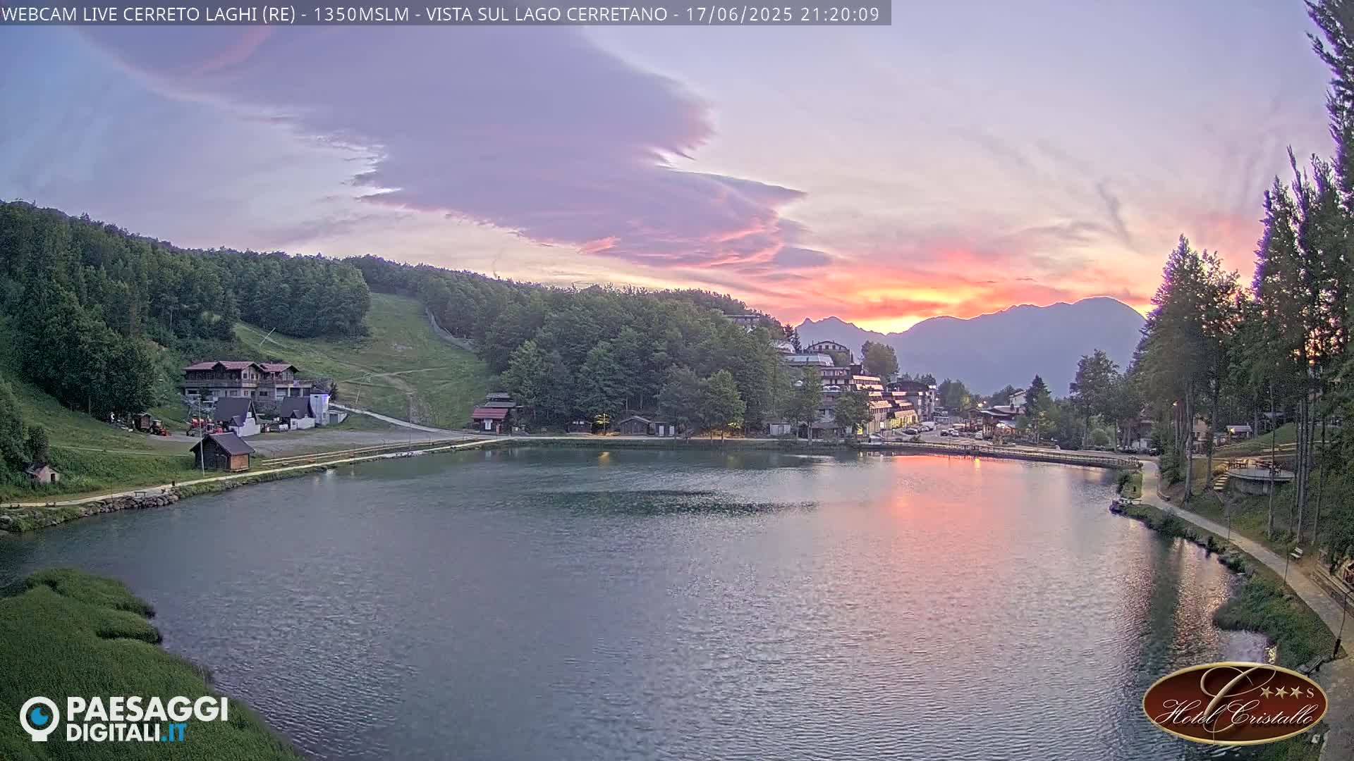 A calm lake reflects a sunset over a mountain range and a village nestled in a wooded area.