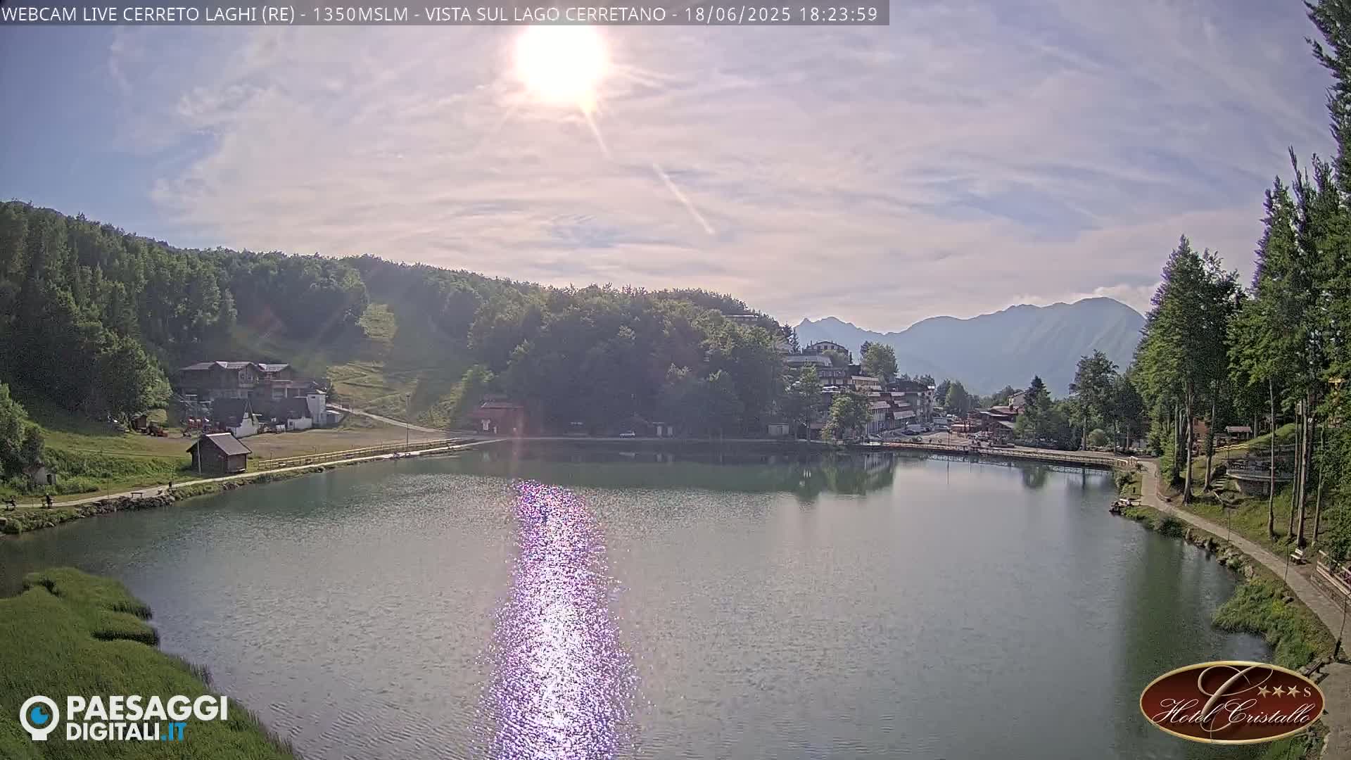 A calm lake reflects the sun's glare, surrounded by green hills, a small village, and distant mountains under a partly cloudy sky.