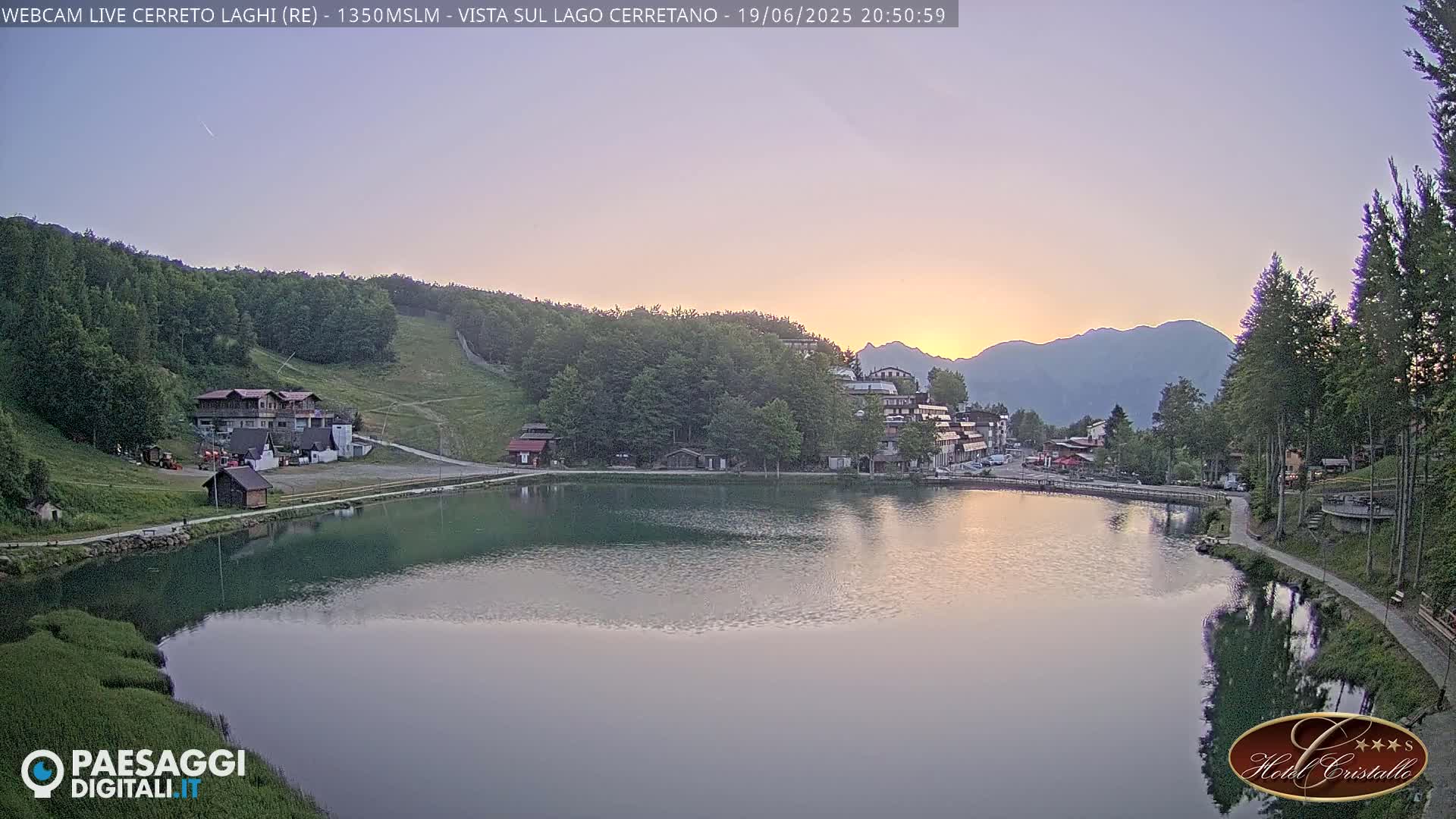A calm lake reflects the sunset over a mountain village nestled in a green valley.