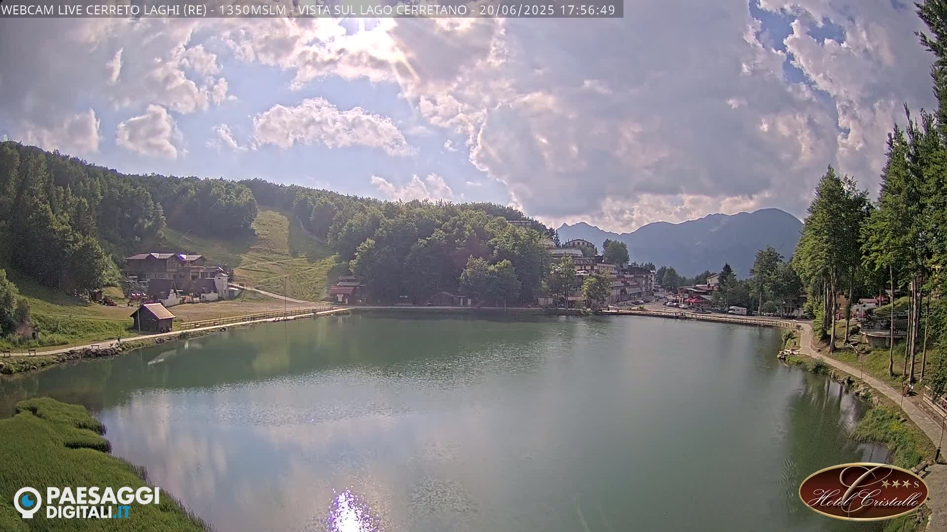 A partly cloudy day reveals a calm lake nestled in a mountainous region, with buildings and trees surrounding the shoreline.