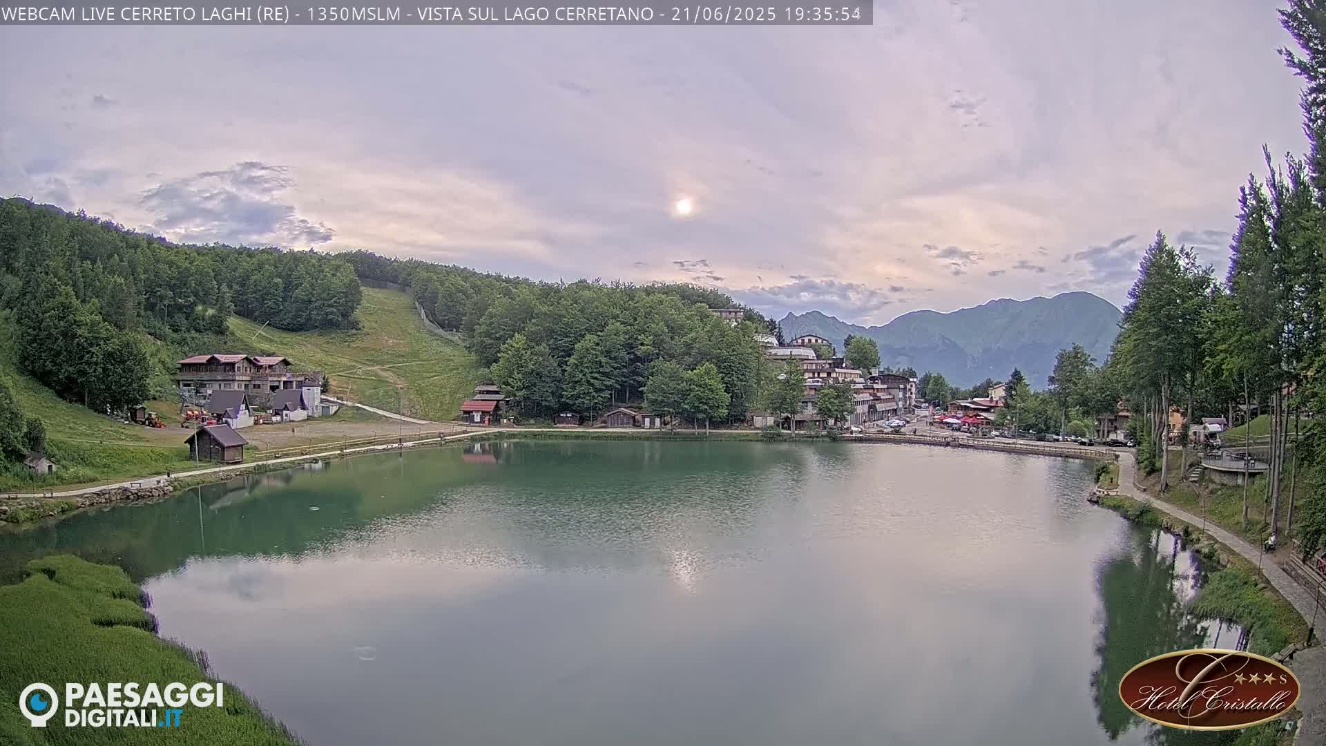 A calm lake reflects the setting sun and surrounding mountains under a partly cloudy sky, with buildings and trees lining its shores.