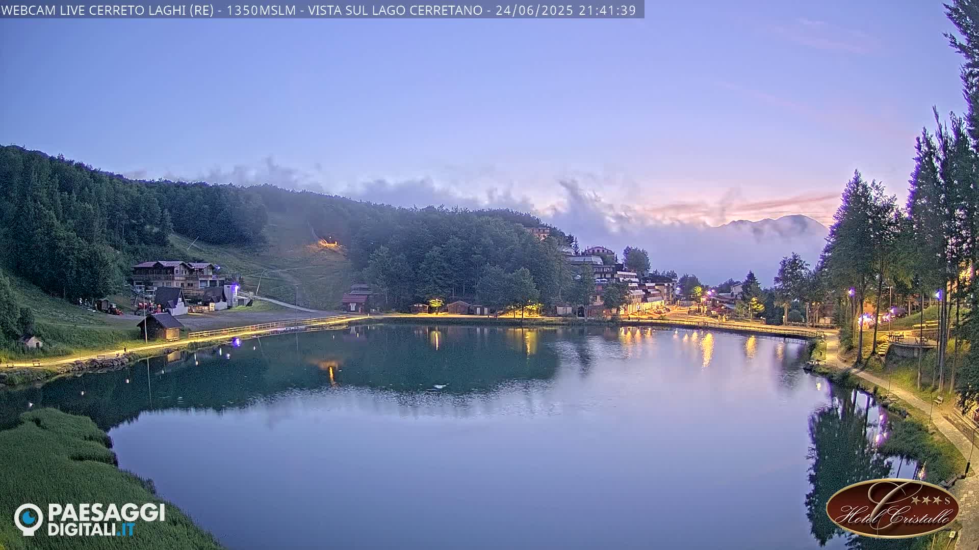A calm lake reflects the lights of a village nestled in a forest at twilight under a mostly clear sky.