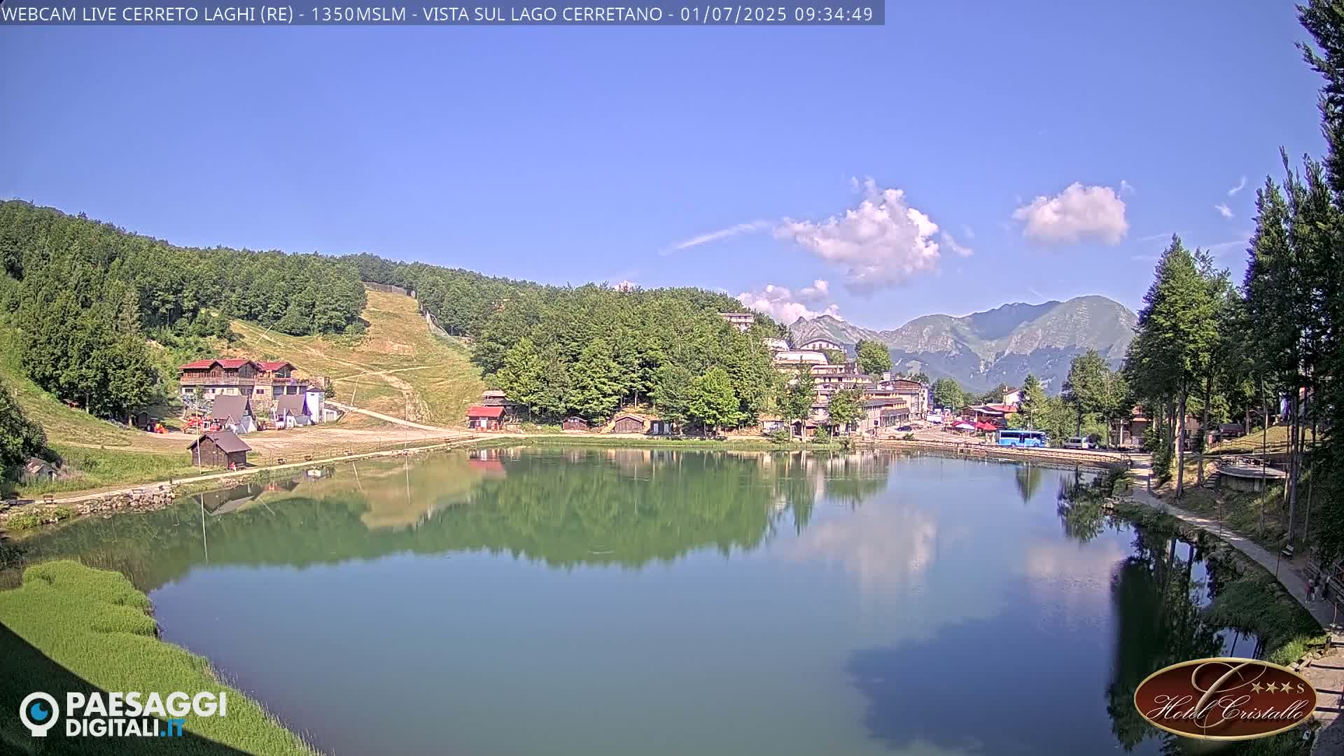 A calm lake reflects a mountain village and surrounding hills under a mostly sunny sky with a few scattered clouds.