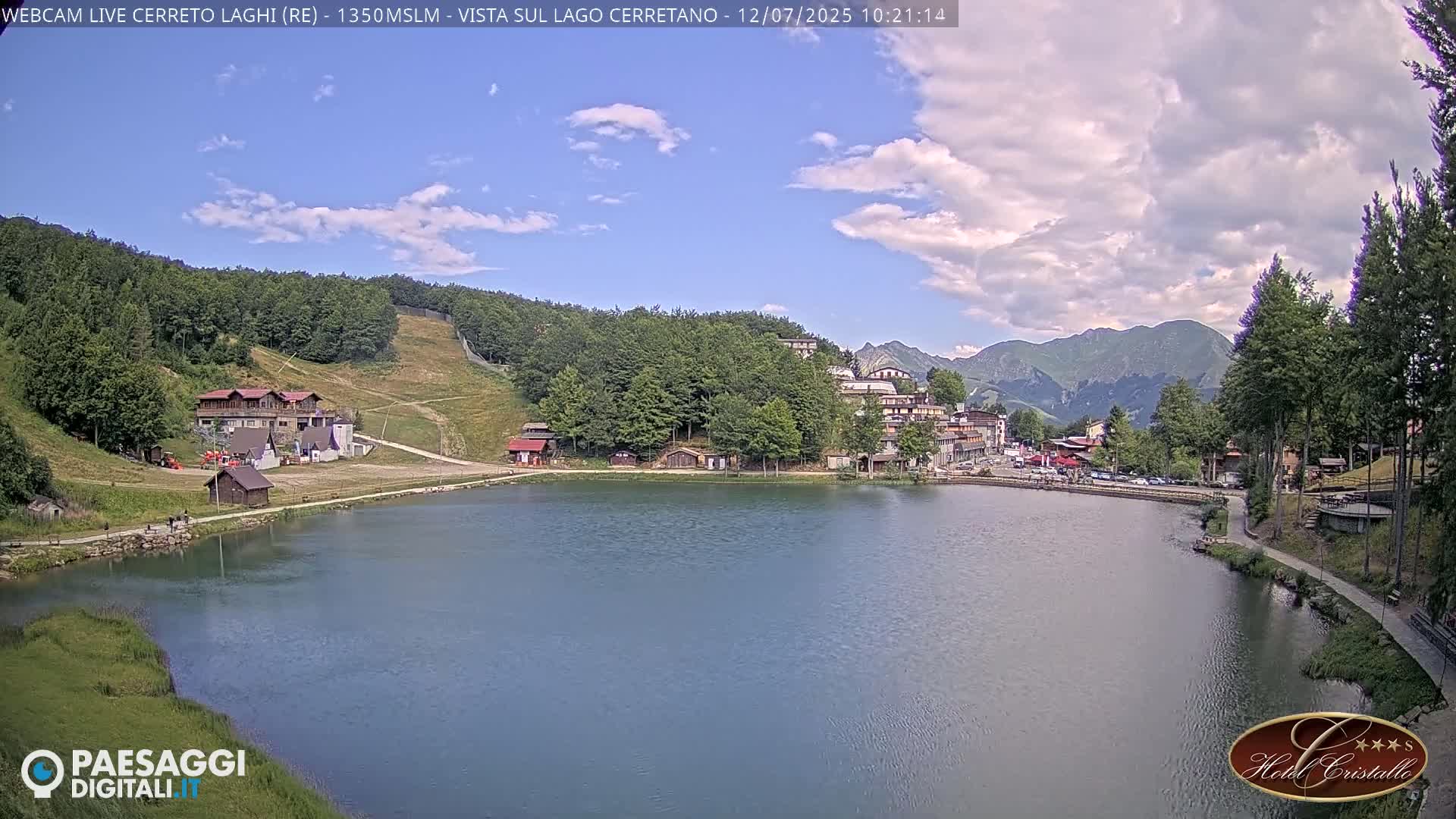 A calm lake is nestled in a valley surrounded by green hills, trees, and mountains under a partly cloudy sky.