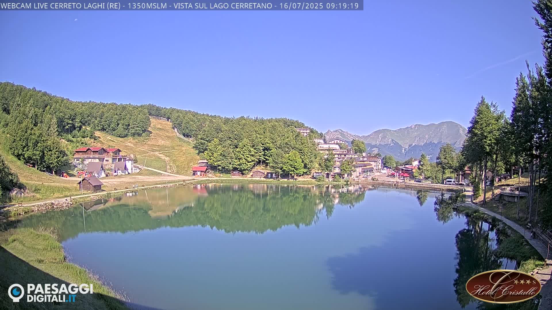 A calm lake reflects buildings and mountains under a clear blue sky.