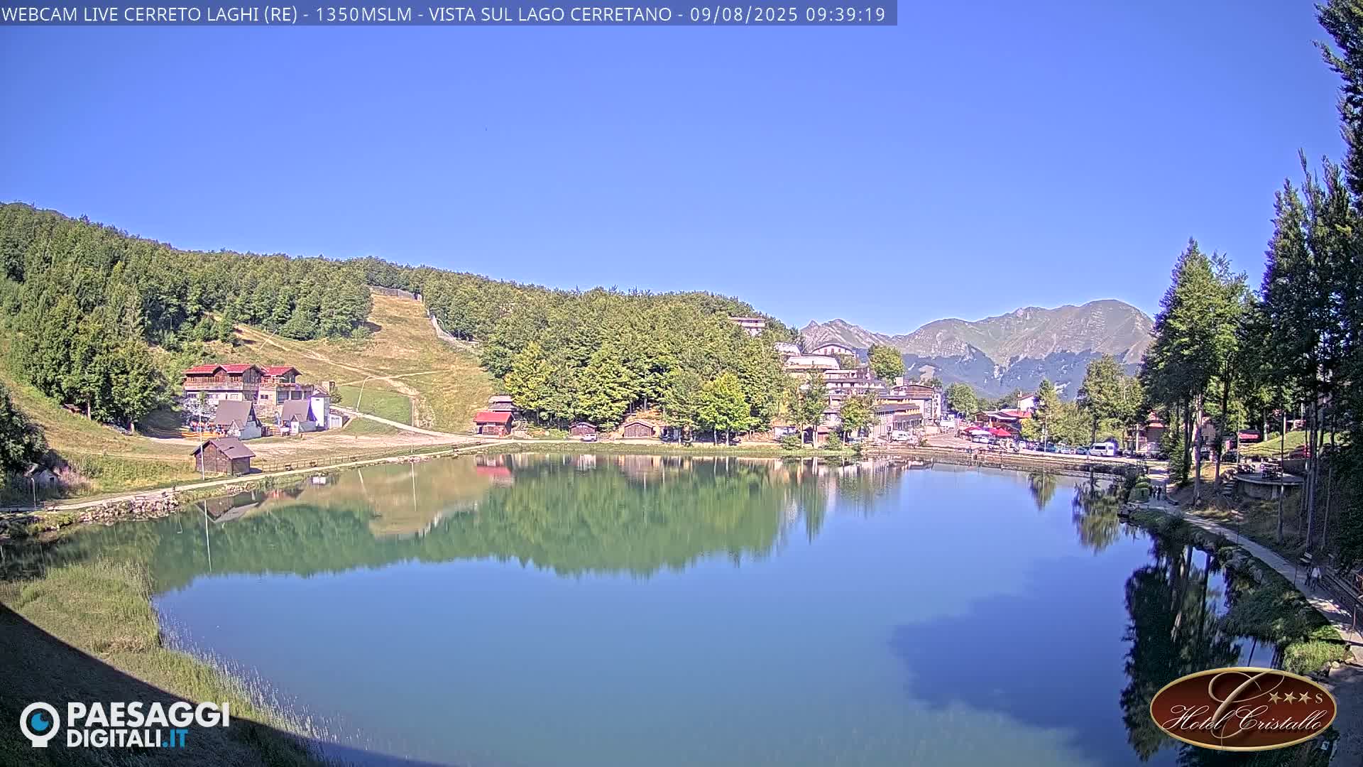 A calm lake reflects a mountain village and surrounding hills under a clear blue sky.
