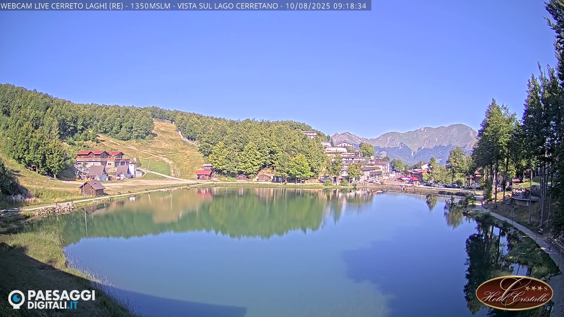 A calm, clear day reveals a mountain lake reflecting buildings and trees, with mountains in the background.