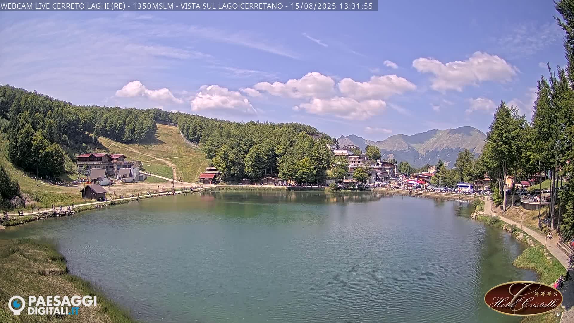 A calm lake reflects a village nestled among green hills and mountains under a clear blue sky.