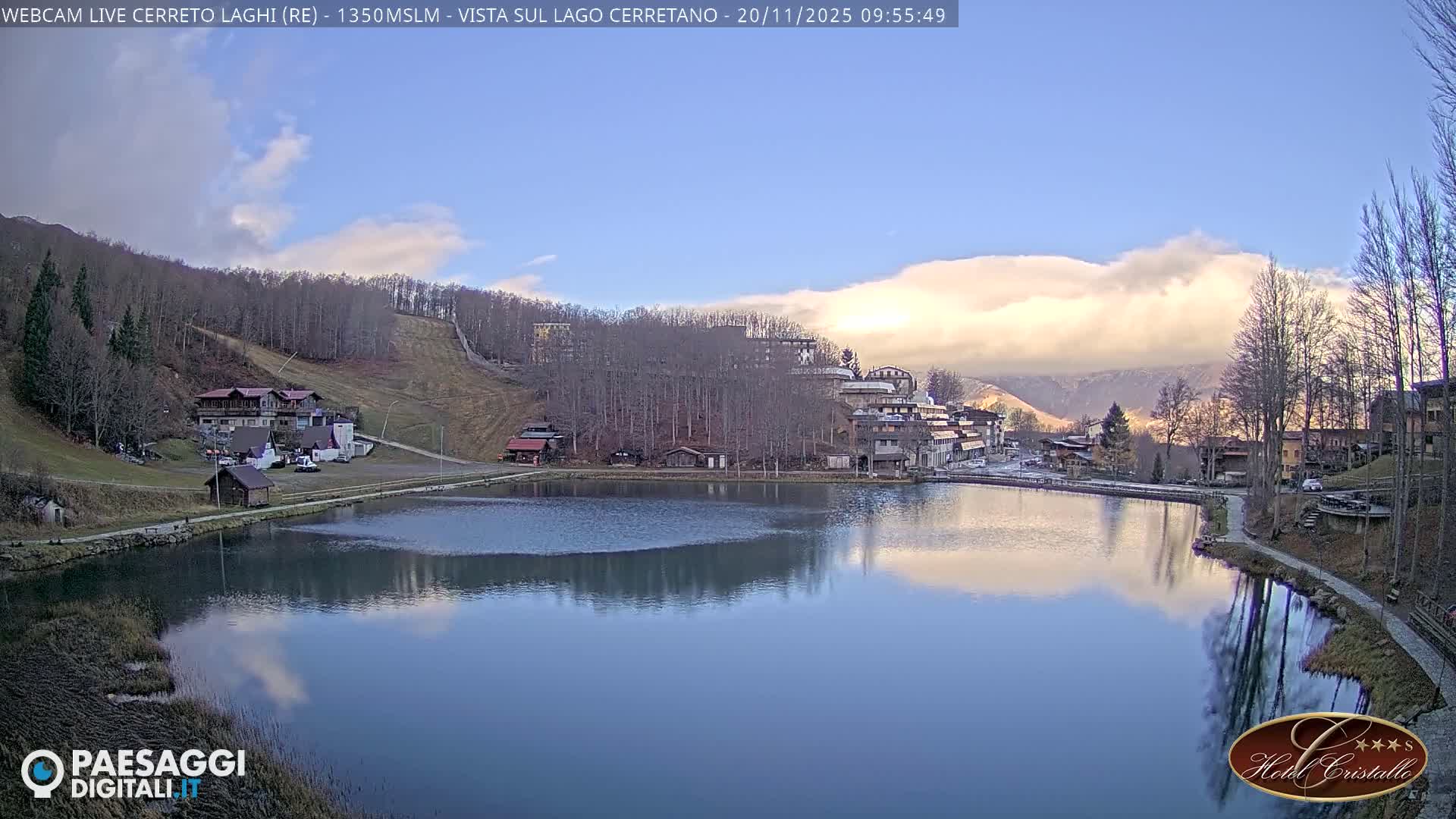 A tranquil mountain village with buildings nestled on forested slopes surrounds a calm lake, reflecting a partly cloudy sky on a cool, crisp day.