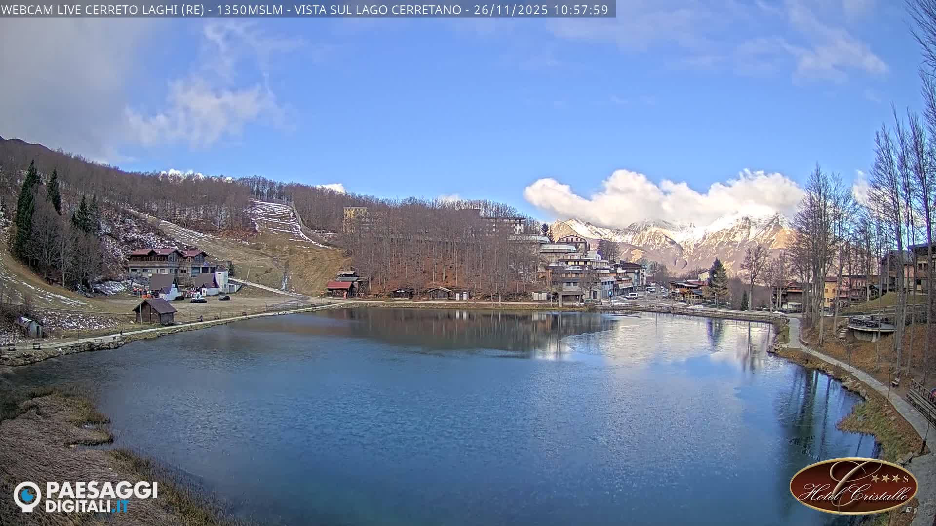 A clear, sunny day reveals a serene mountain village nestled around a reflective lake, with bare-treed hillsides and distant snow-capped mountains under a blue sky adorned with scattered white clouds.