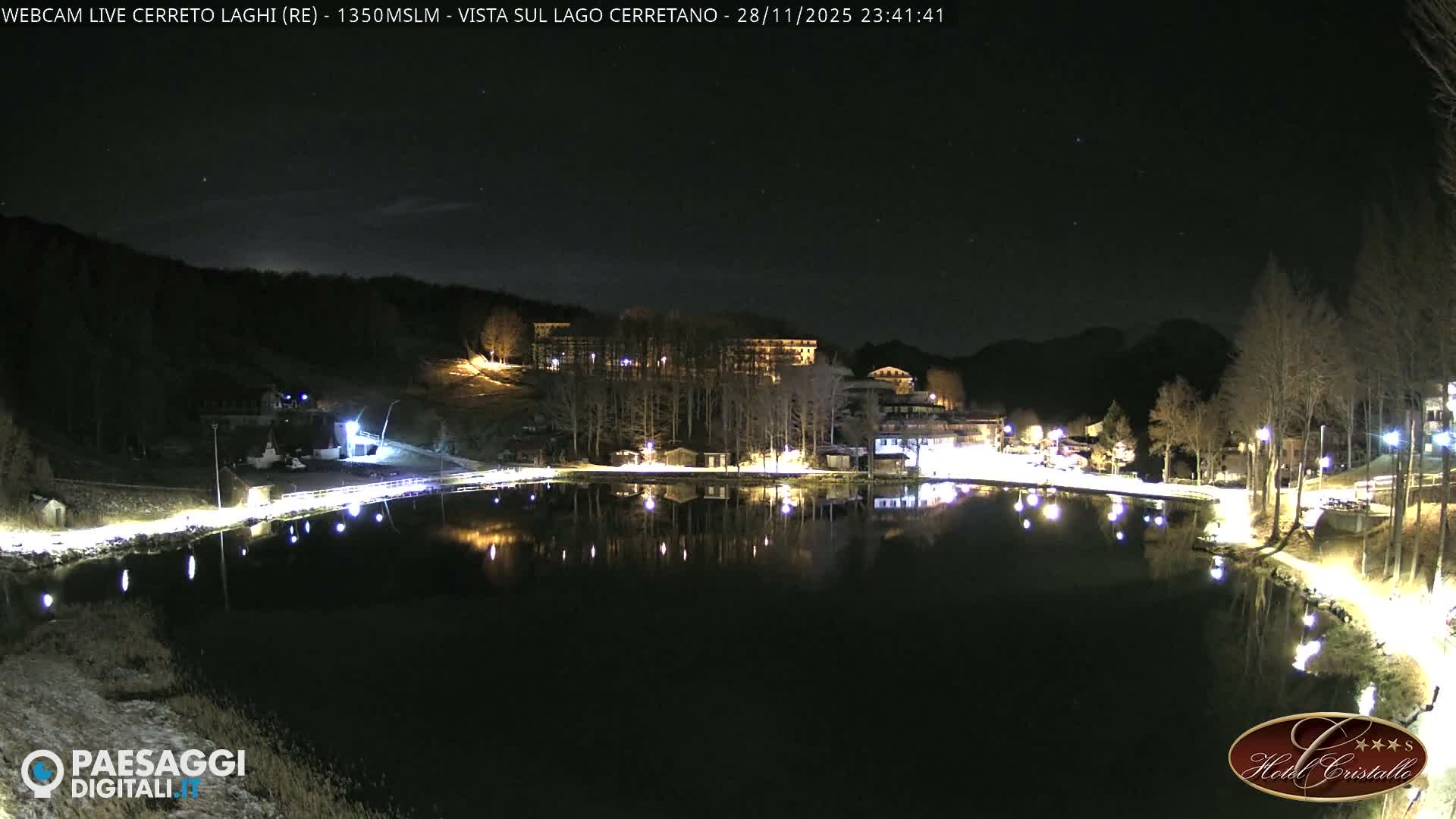 A clear, star-filled night illuminates a serene mountain lake reflecting the bright lights of surrounding buildings and a lakeside path, with bare trees and distant dark mountains.