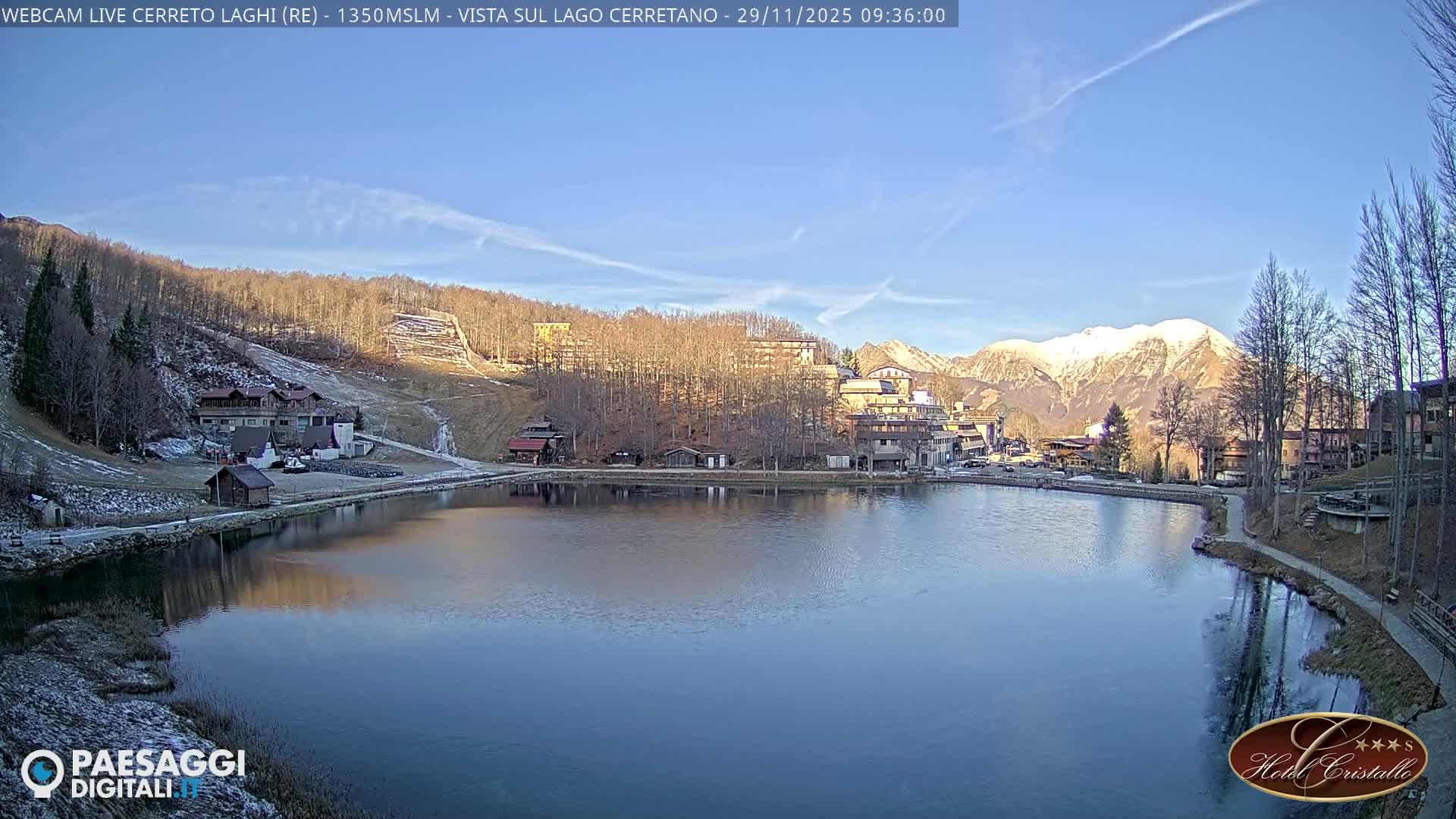 A serene winter landscape features a still, partially frozen lake reflecting a clear blue sky, bordered by a village with buildings and bare trees, set against a backdrop of snow-capped mountains.