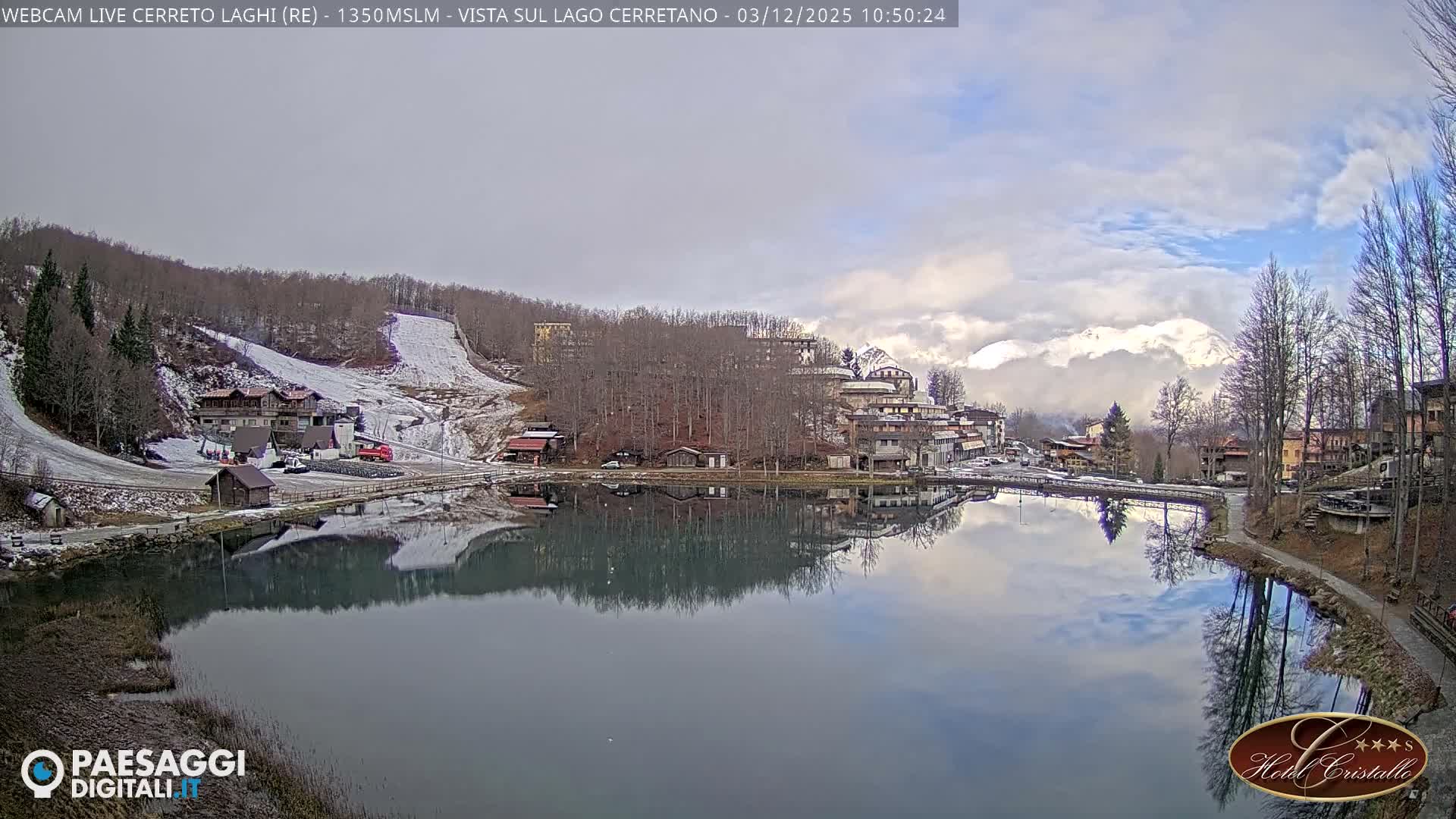 A tranquil alpine lake reflects a partially cloudy sky and a village of buildings and bare trees nestled on snow-dusted hillsides, all beneath distant, cloud-shrouded snowy mountain peaks on a cool, partially cloudy day.