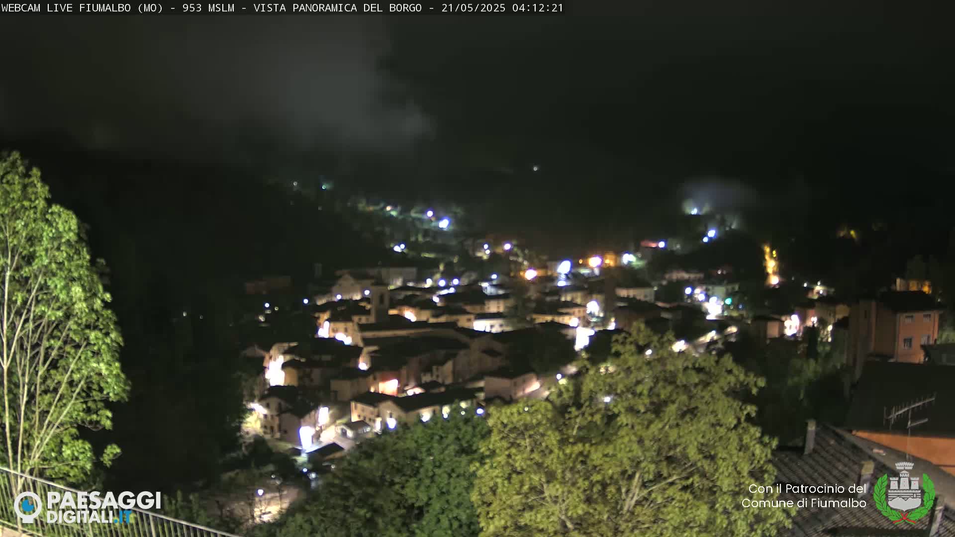 A nighttime, panoramic view of a small town nestled among hills and trees, illuminated by streetlights.