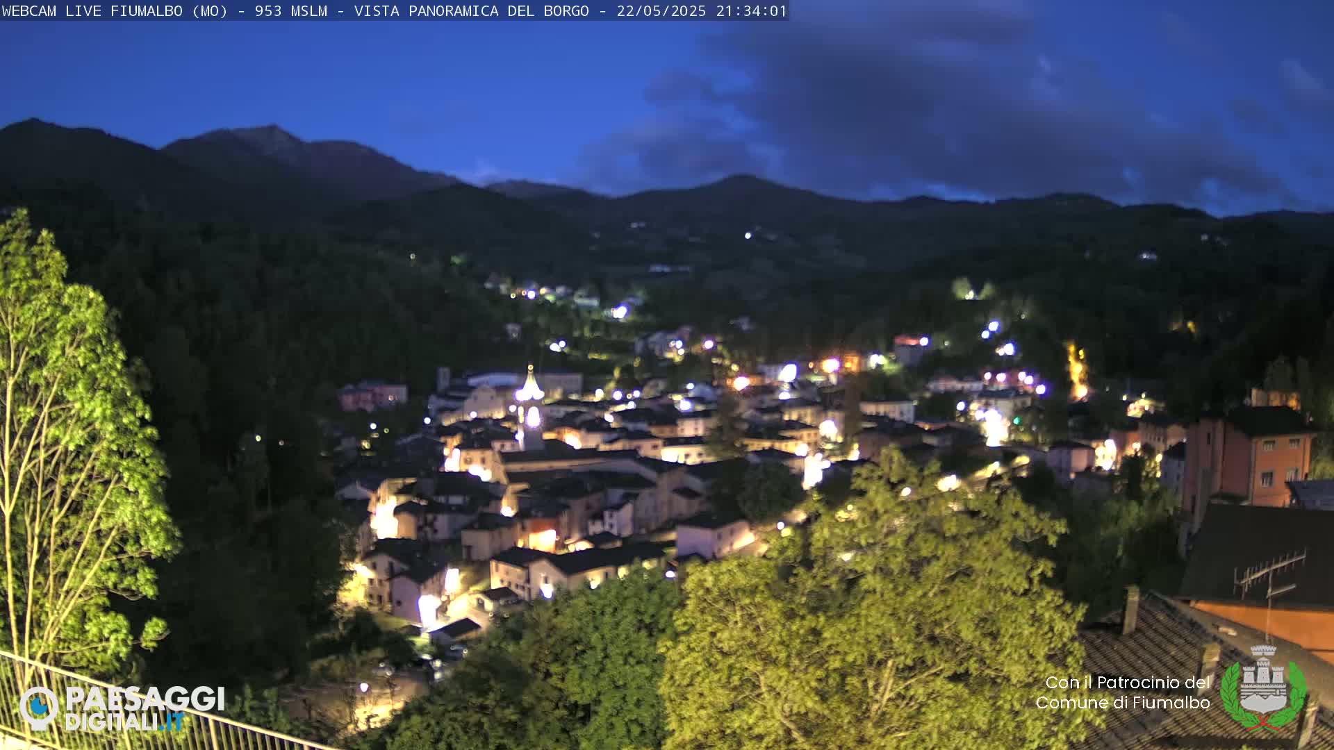 A nighttime panoramic view of a small village nestled in a valley between mountains under a partly cloudy sky.