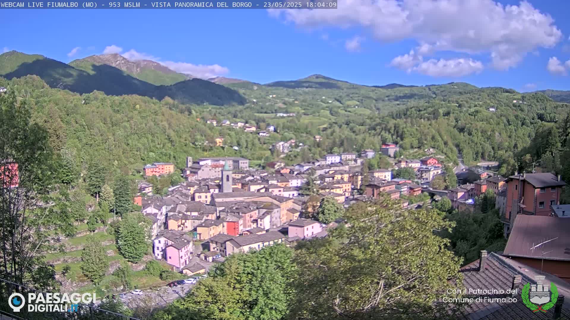 A picturesque village nestled in a valley surrounded by lush green hills and mountains under a partly cloudy sky.