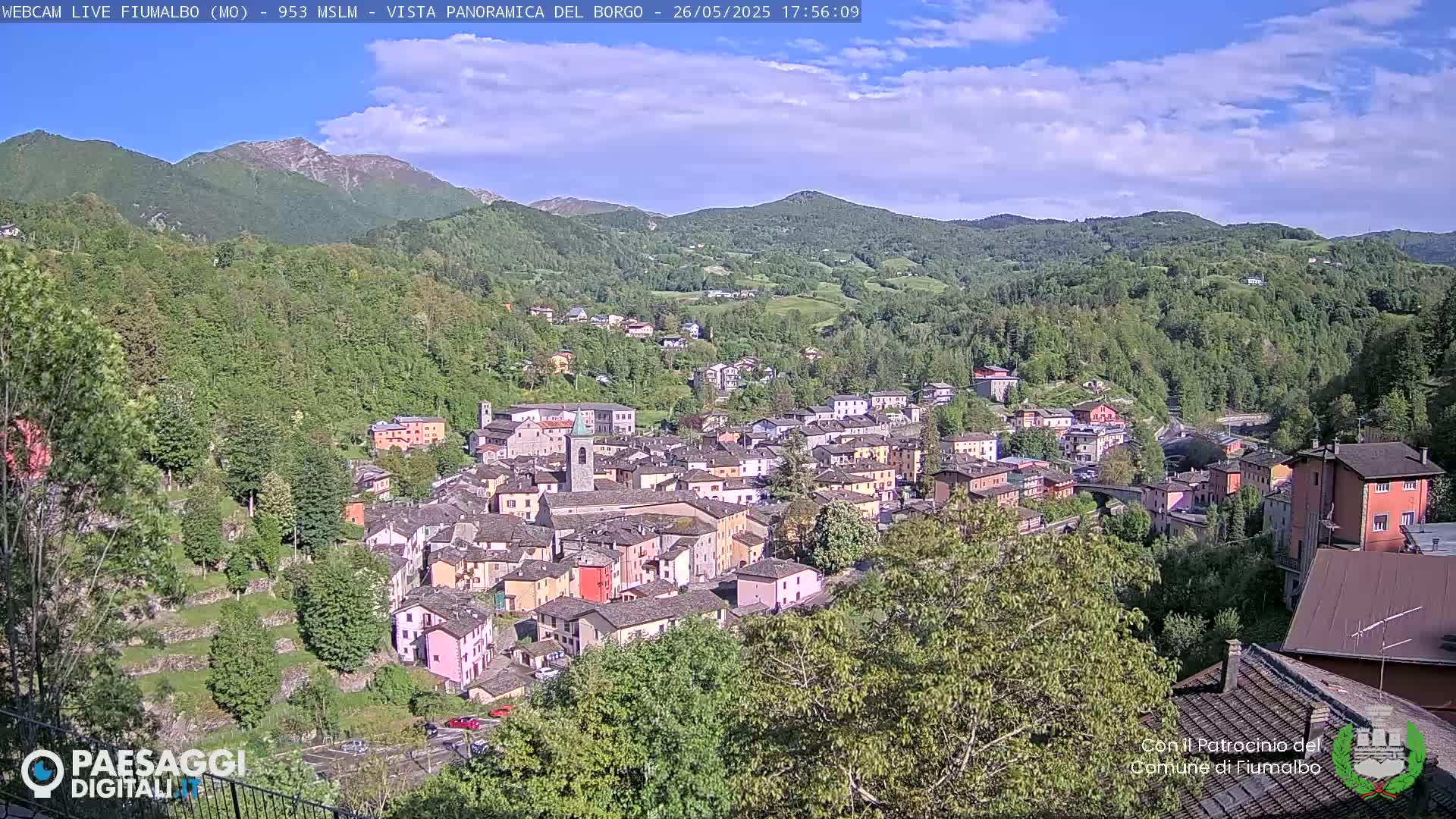 A picturesque village nestled in a valley surrounded by lush green hills and mountains under a mostly sunny sky.