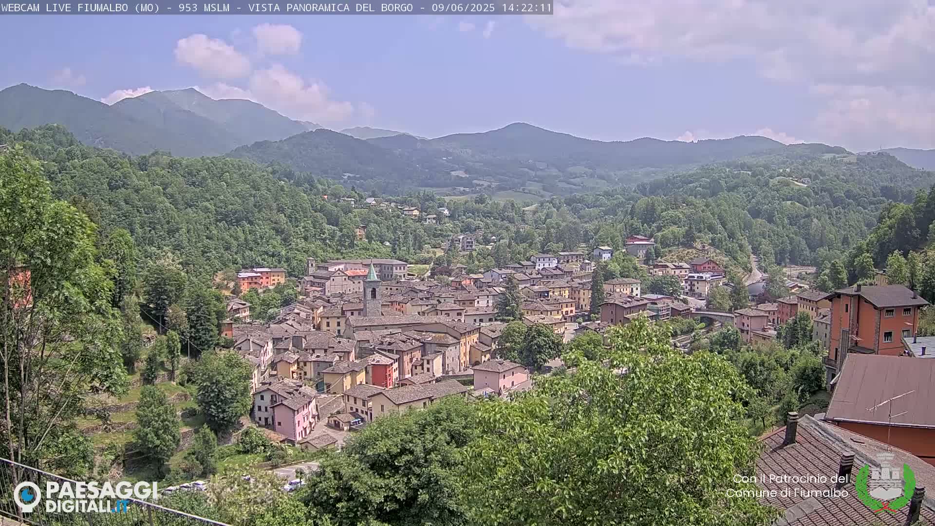 A small town nestled in a valley, surrounded by lush green hills and mountains under a partly cloudy sky.