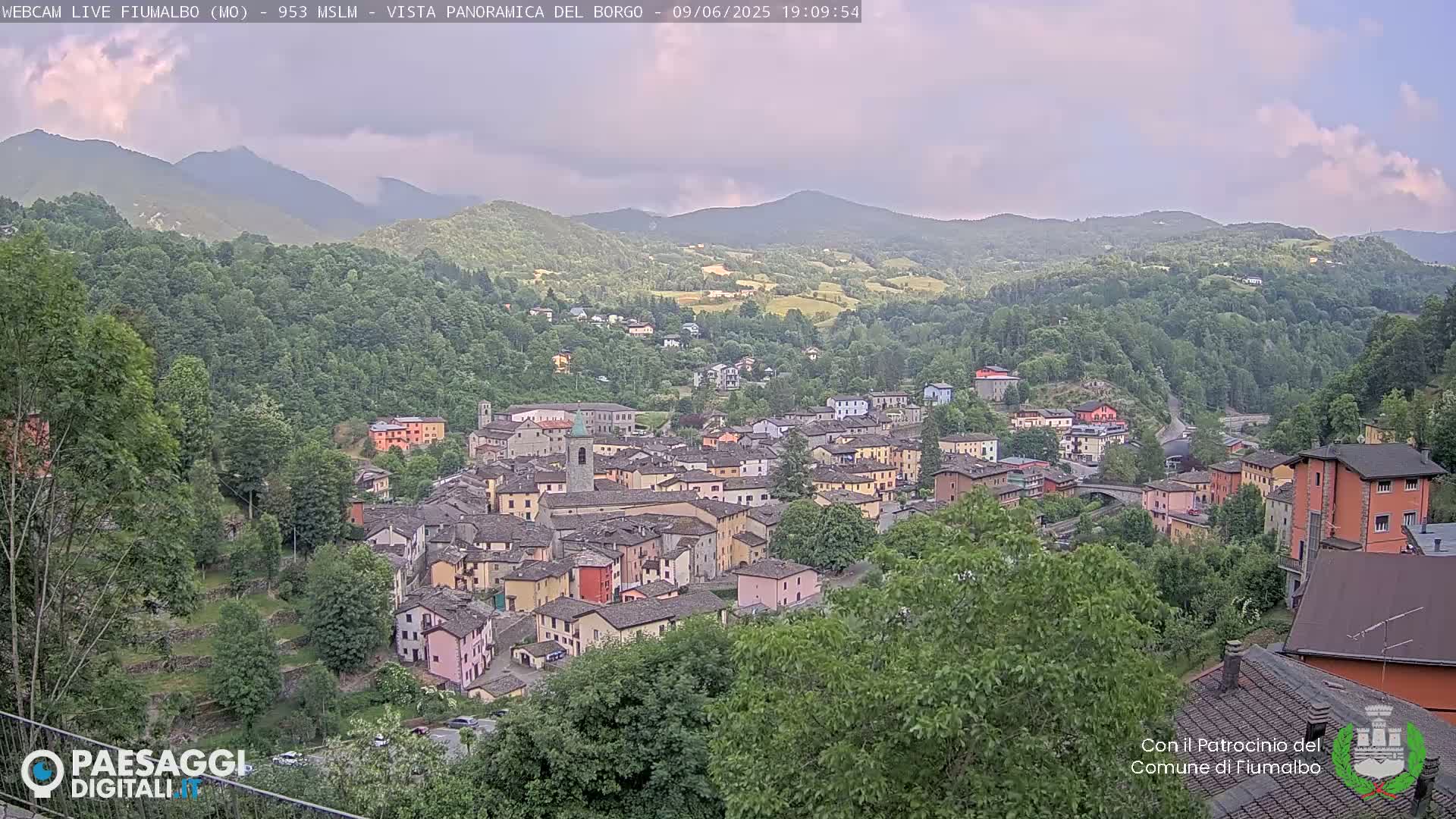 A picturesque village nestled in a valley surrounded by green hills and mountains under a partly cloudy sky.