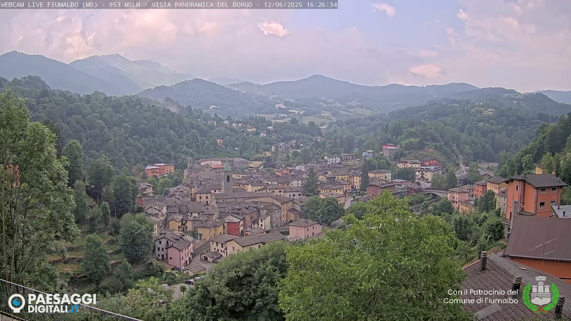 A hazy, slightly overcast view shows a small town nestled in a valley surrounded by lush green hills and mountains.
