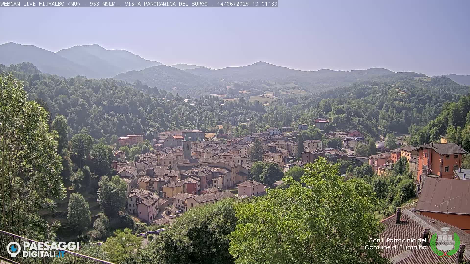 A small town nestled in a valley, surrounded by lush green hills and mountains under a clear, sunny sky.