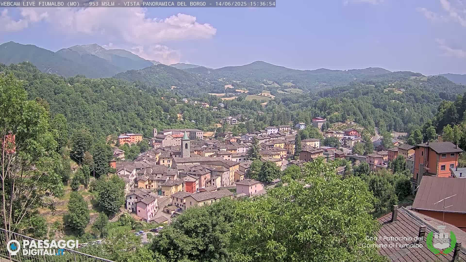 A quaint village nestled in a valley, surrounded by lush green hills and mountains under a mostly clear sky.