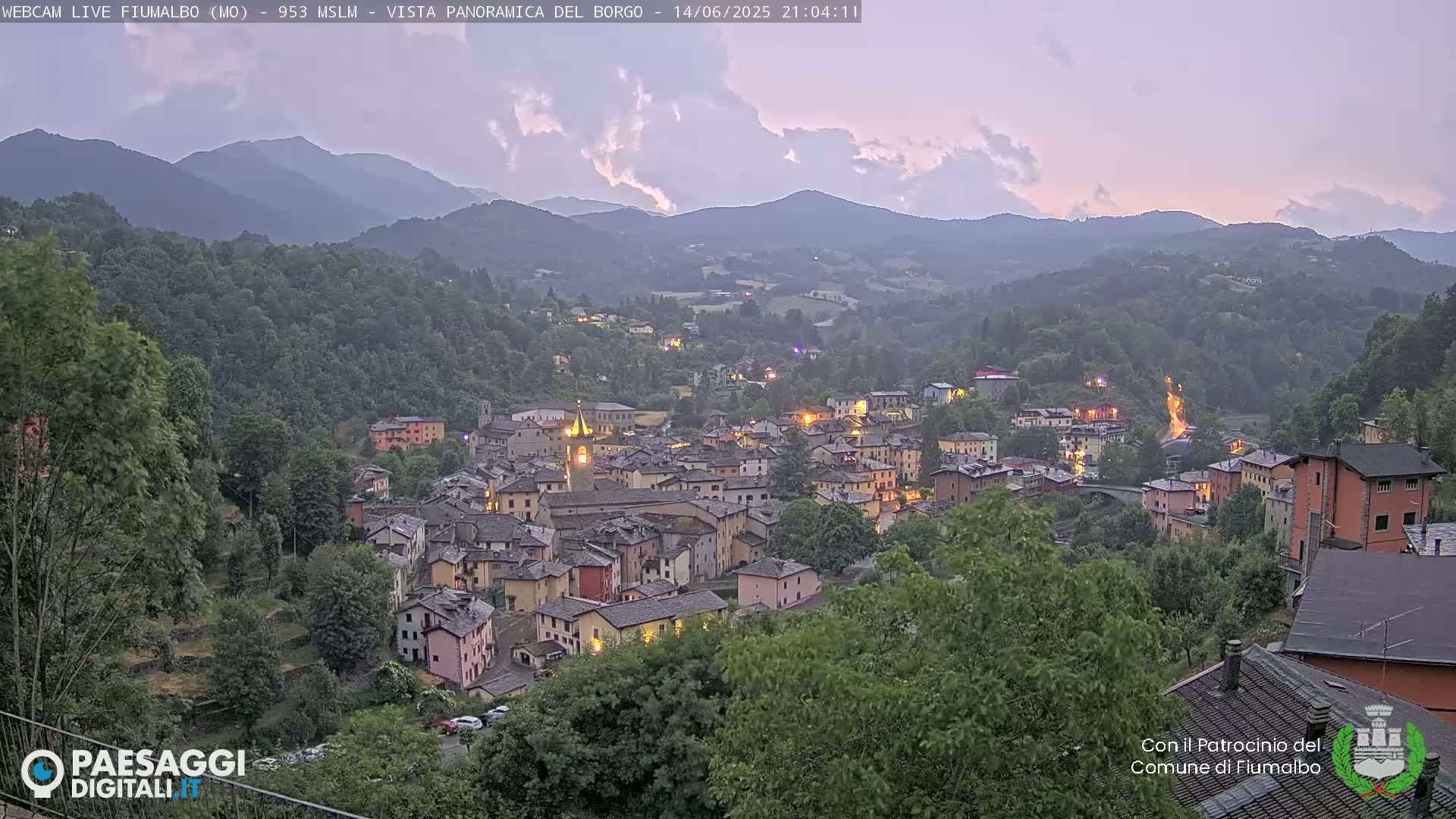 A quaint village nestled in a valley, surrounded by lush green hills and mountains under a mostly clear sky.